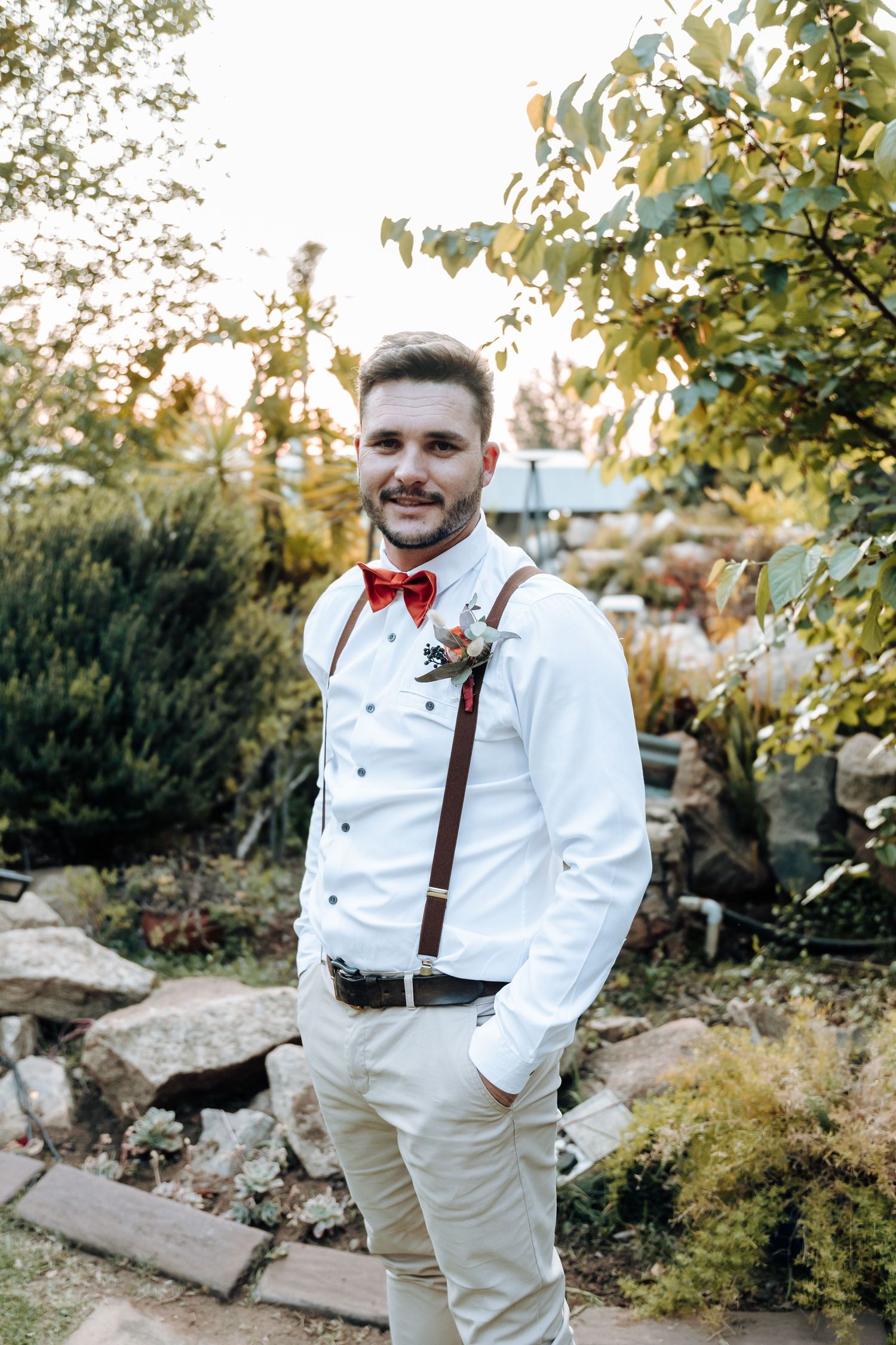 Groomsman wearing white shirt, red bow tie and brown suspenders poses in garden setting.