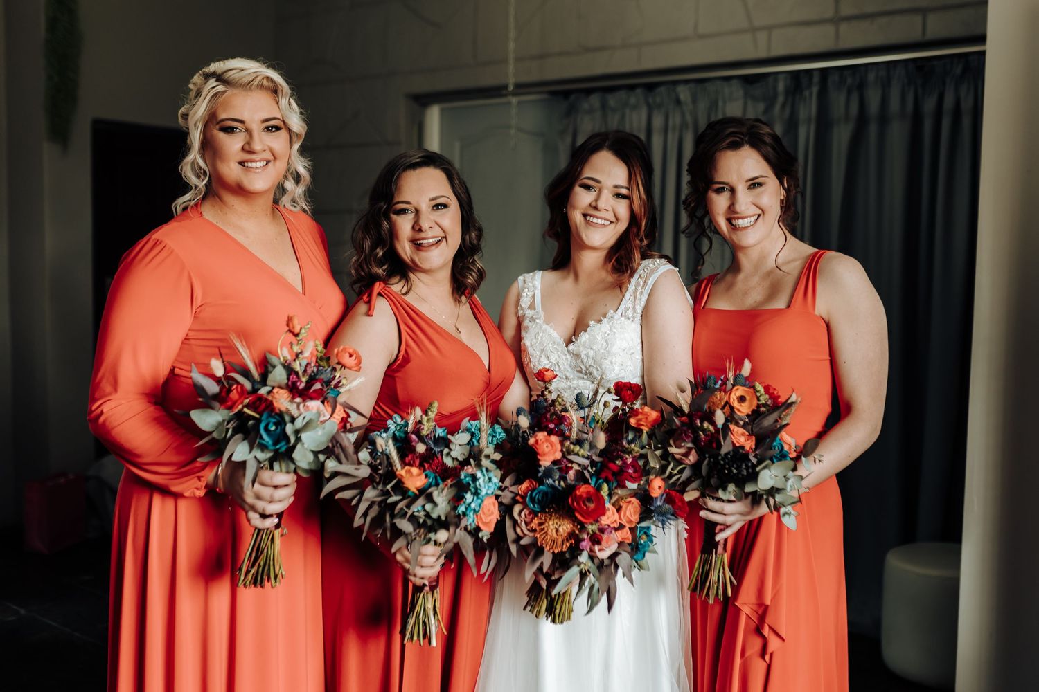 Bridal party wearing coral orange dresses hold vibrant bouquets with blue and coral flowers at indoor venue.