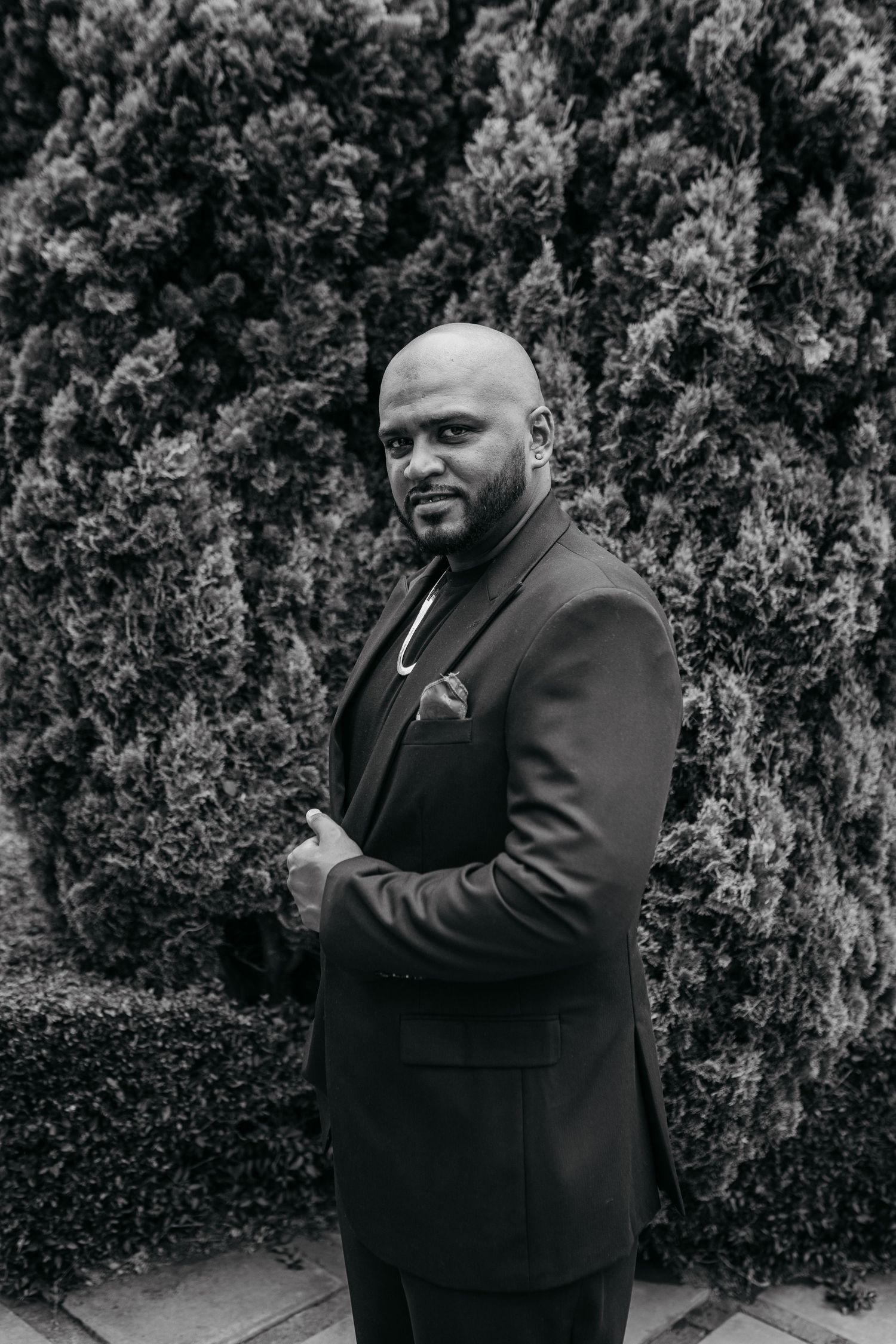 Black and white portrait of person in suit standing against hedge background.