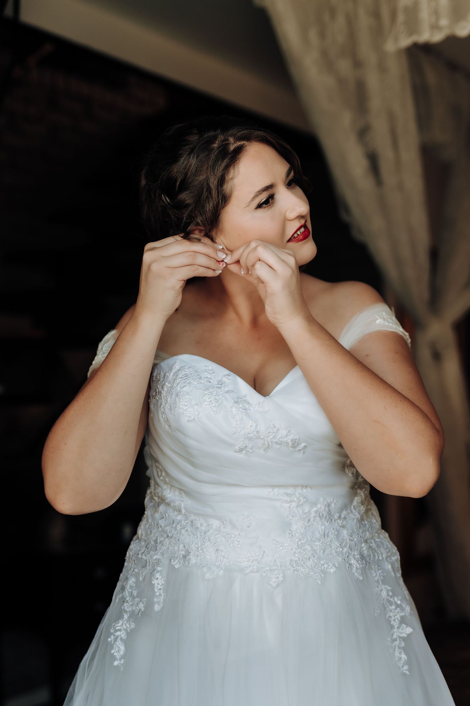 Bride in off-shoulder wedding dress puts on earrings while getting ready.