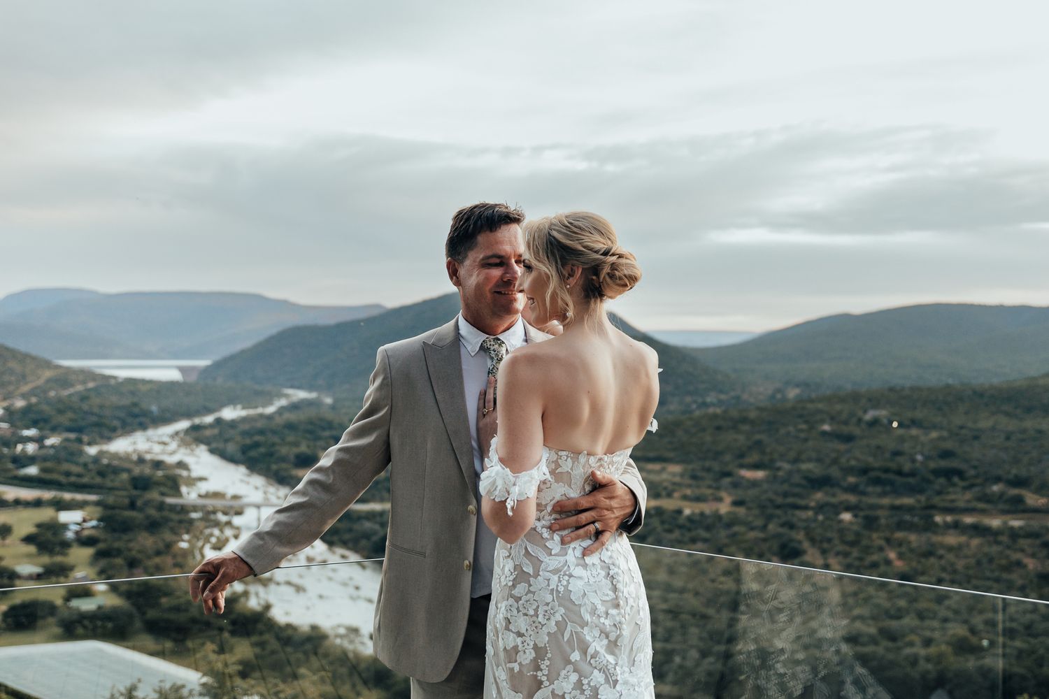 Wedding couple embraces on mountain overlook deck with scenic river valley behind them.
