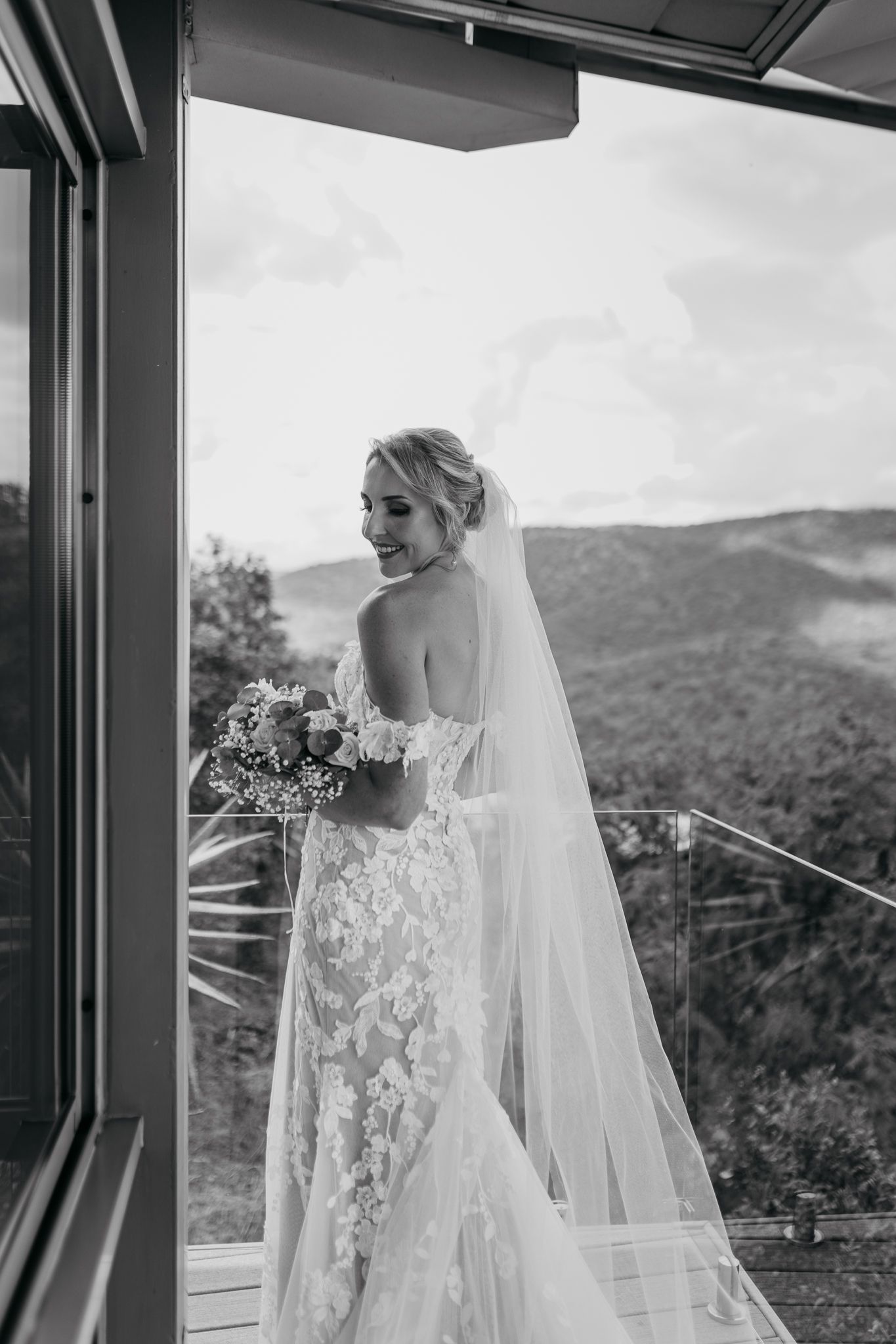 Black and white portrait of bride in lace gown holding bouquet against mountain backdrop.