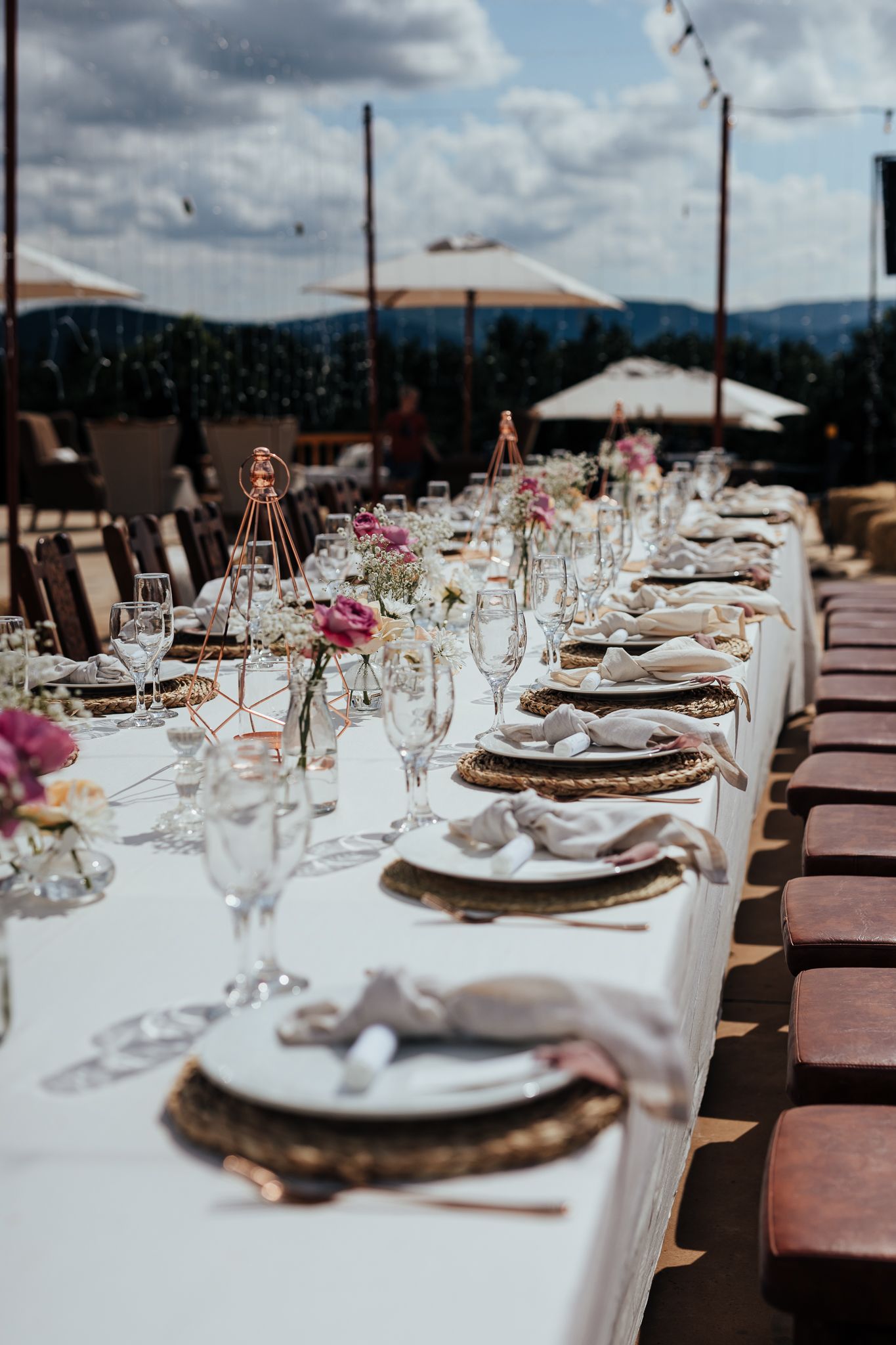 Outdoor wedding reception table with woven chargers and pink floral centerpieces against mountain backdrop.