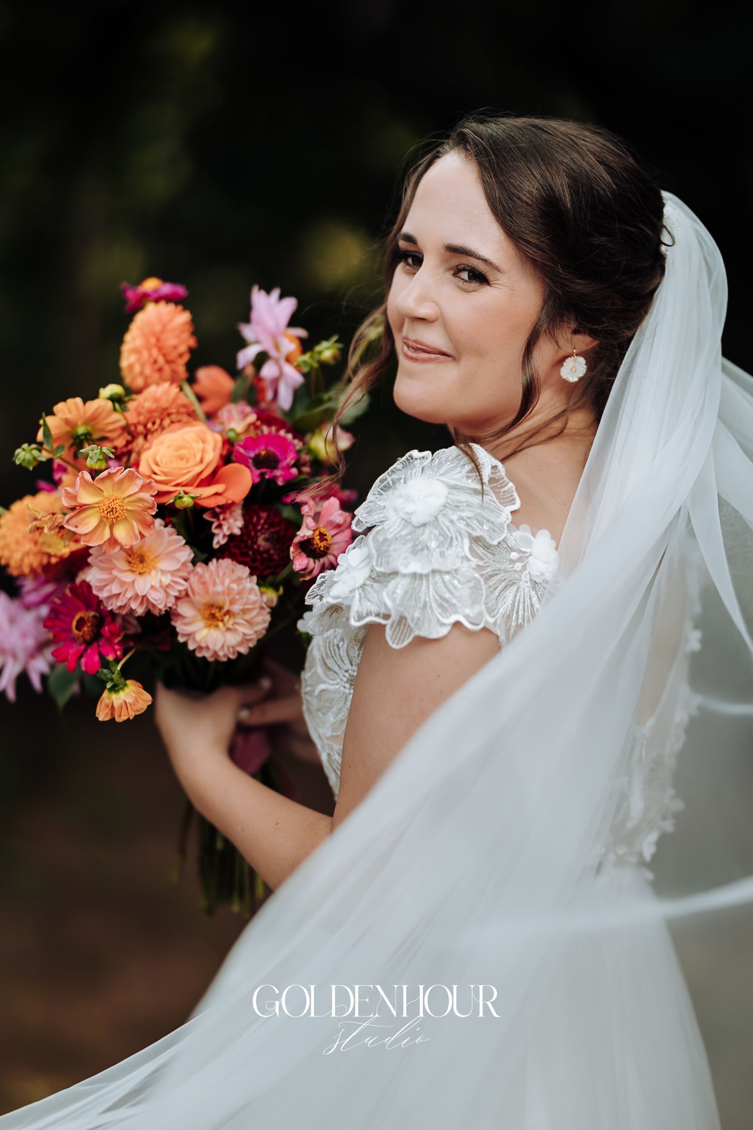 Bride in a lace gown holding a vibrant pink and orange bouquet with veil flowing in the breeze.