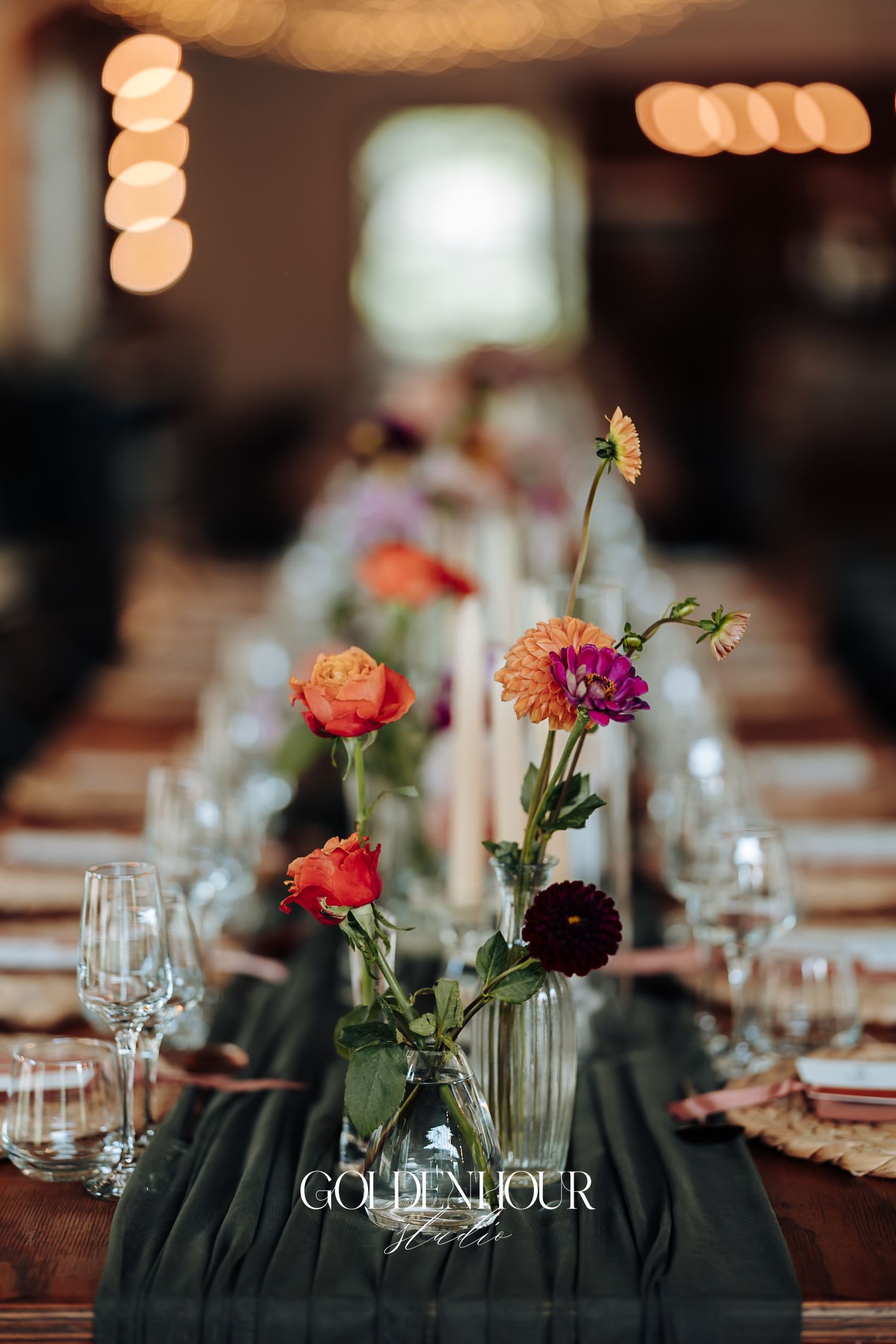 Elegant wedding table centerpiece with colorful wildflowers in glass vases on dark runner with candles and glassware.