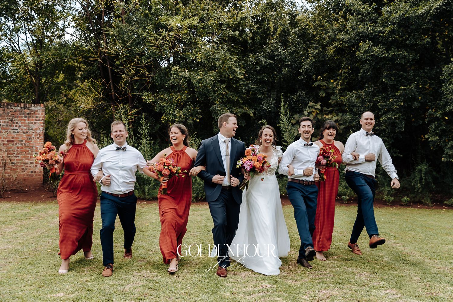 Wedding party in rust and navy walking joyfully across green lawn with bridal bouquets.