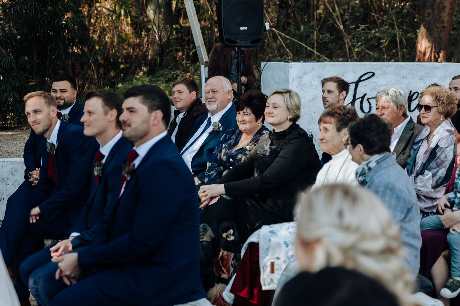 Wedding guests seated outdoors at a garden ceremony, dressed in formal attire including navy suits and elegant dresses.