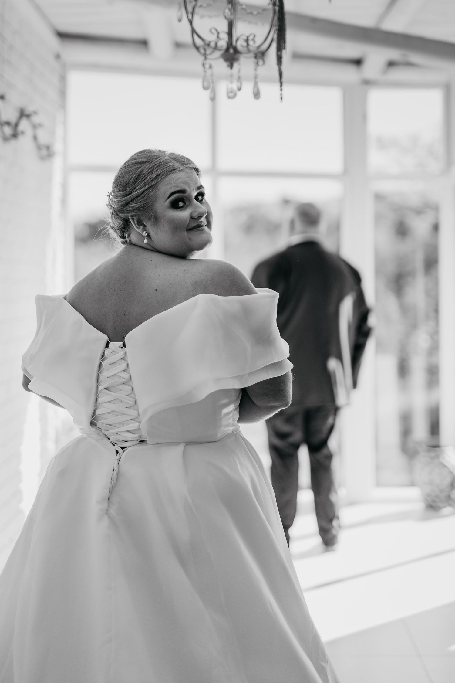 Black and white photo of a bride in an off-shoulder gown with corset back, glancing over shoulder as groom stands nearby.