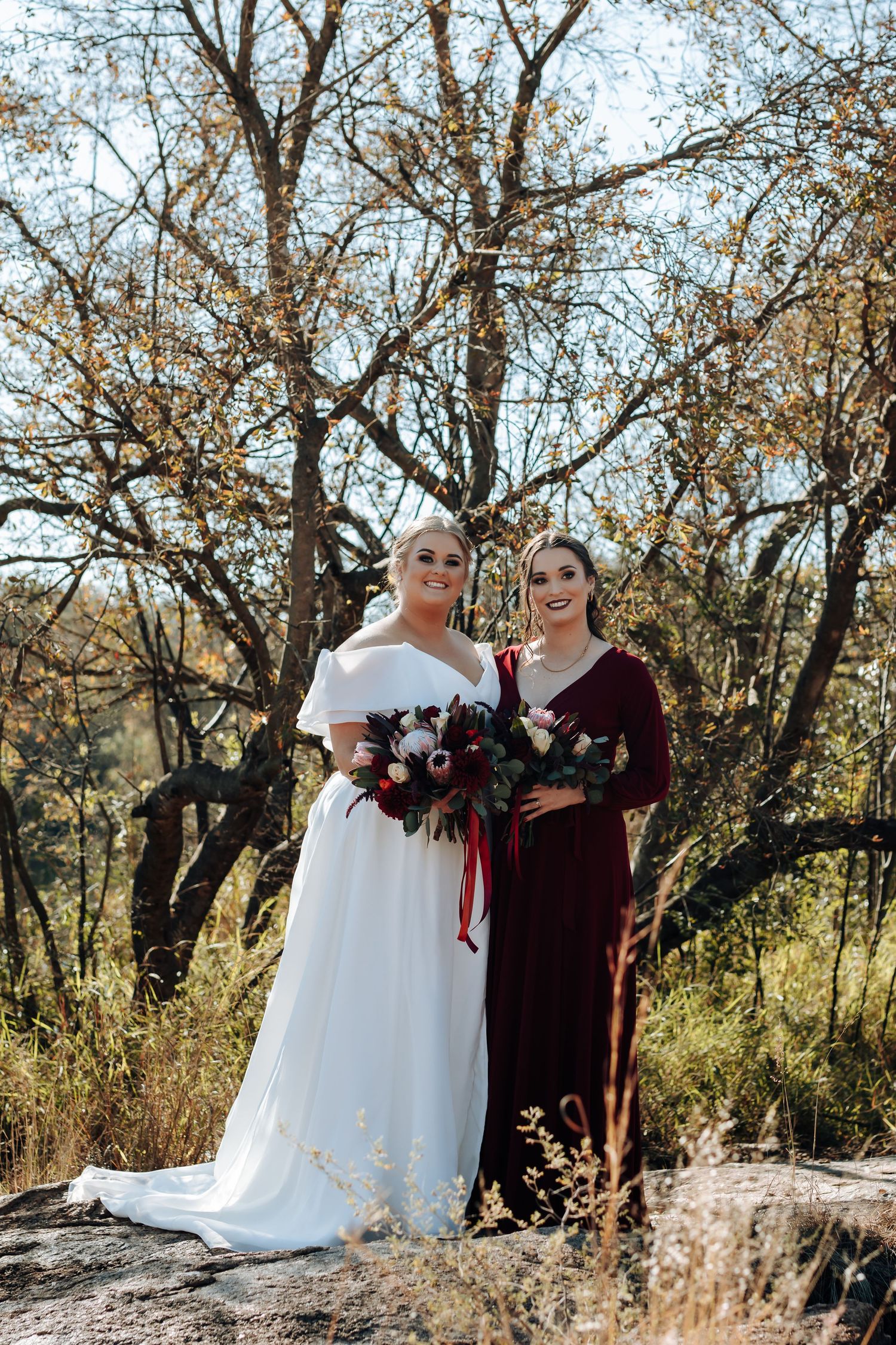 Bride in white gown and bridesmaid in burgundy dress posing outdoors with fall foliage and bouquet.