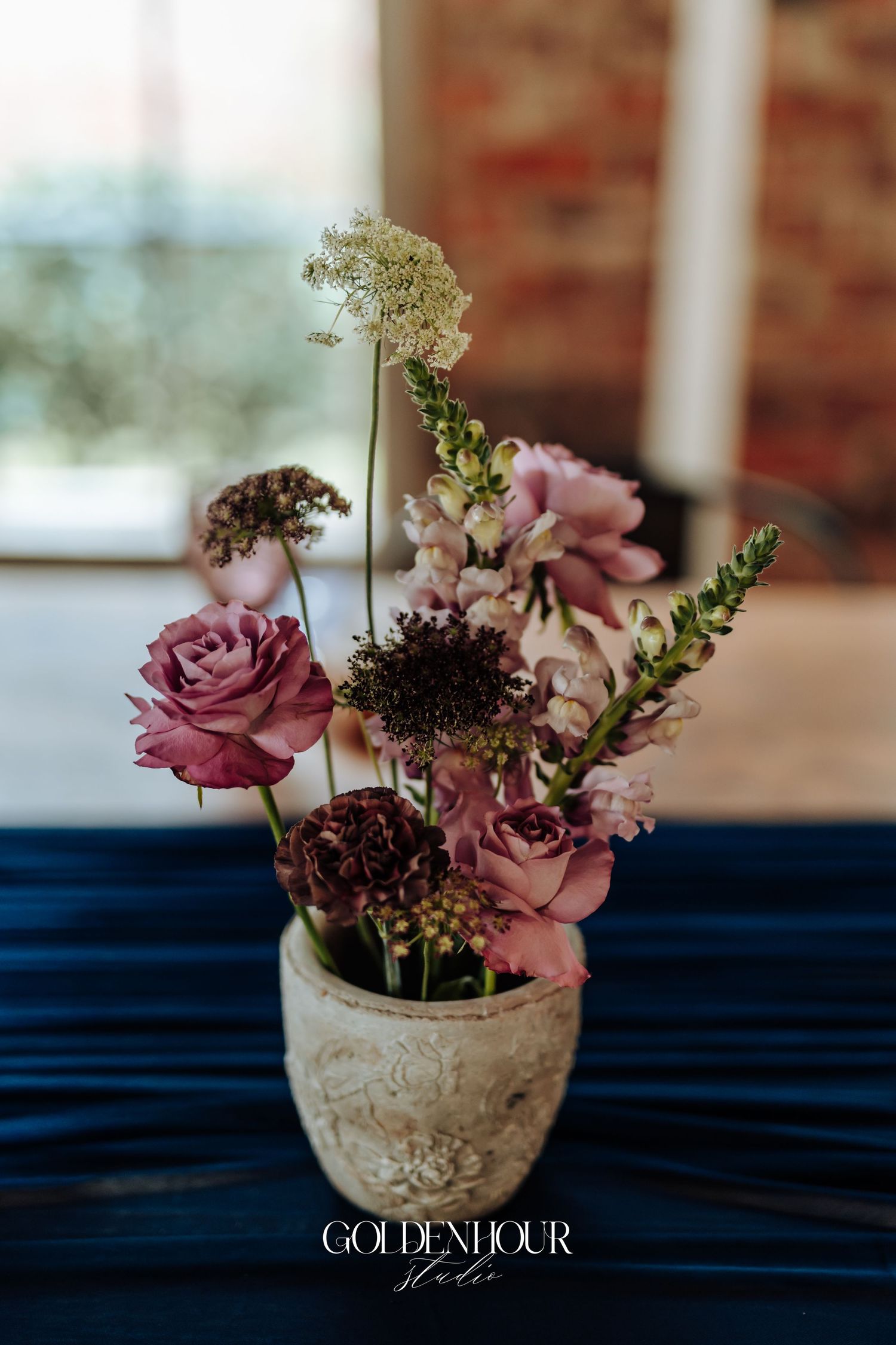 Rustic ceramic vase with pink roses, snapdragons, and wildflowers centerpiece on navy blue satin table.