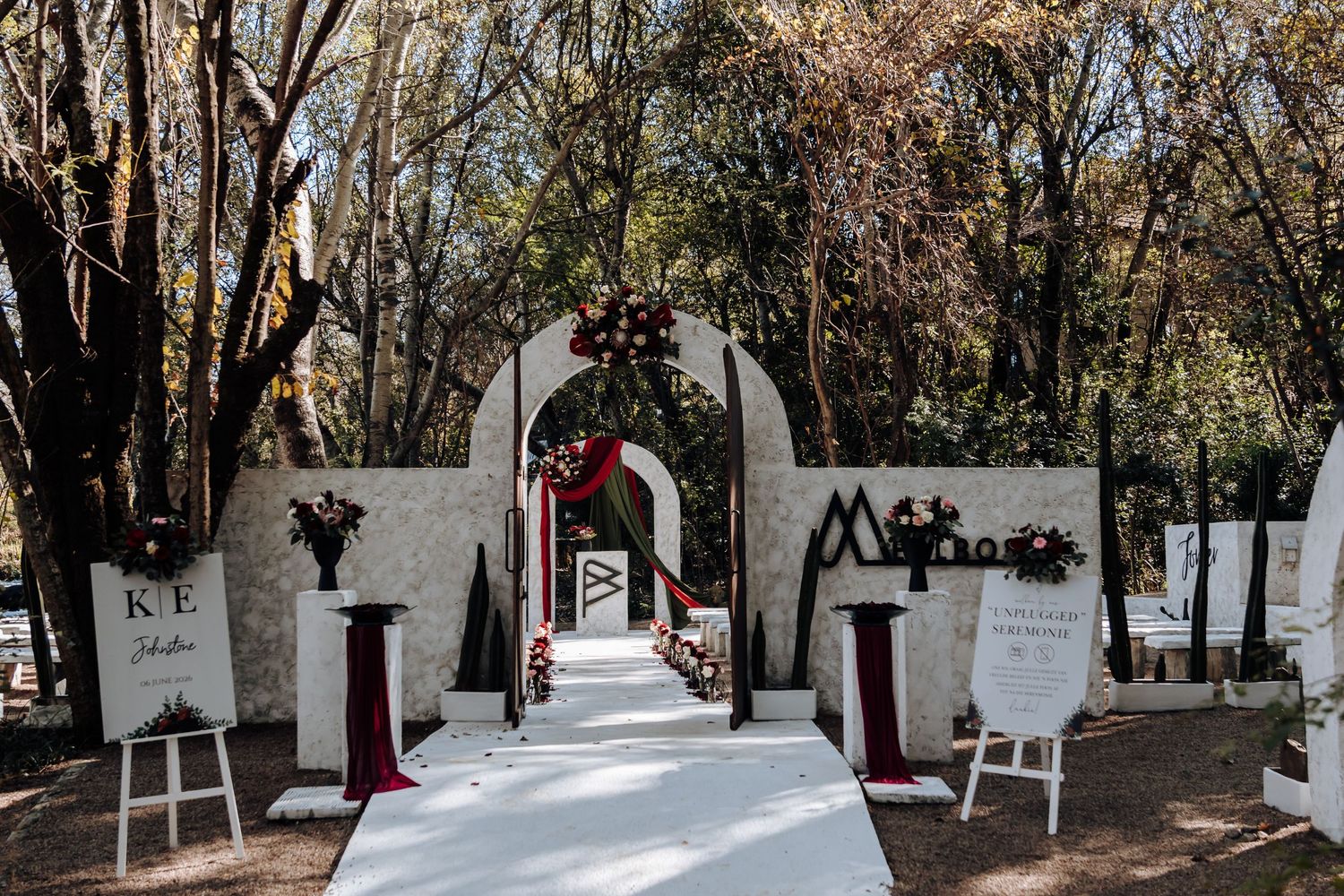 Elegant outdoor wedding ceremony aisle with white arch, red floral accents, and tree-lined backdrop.