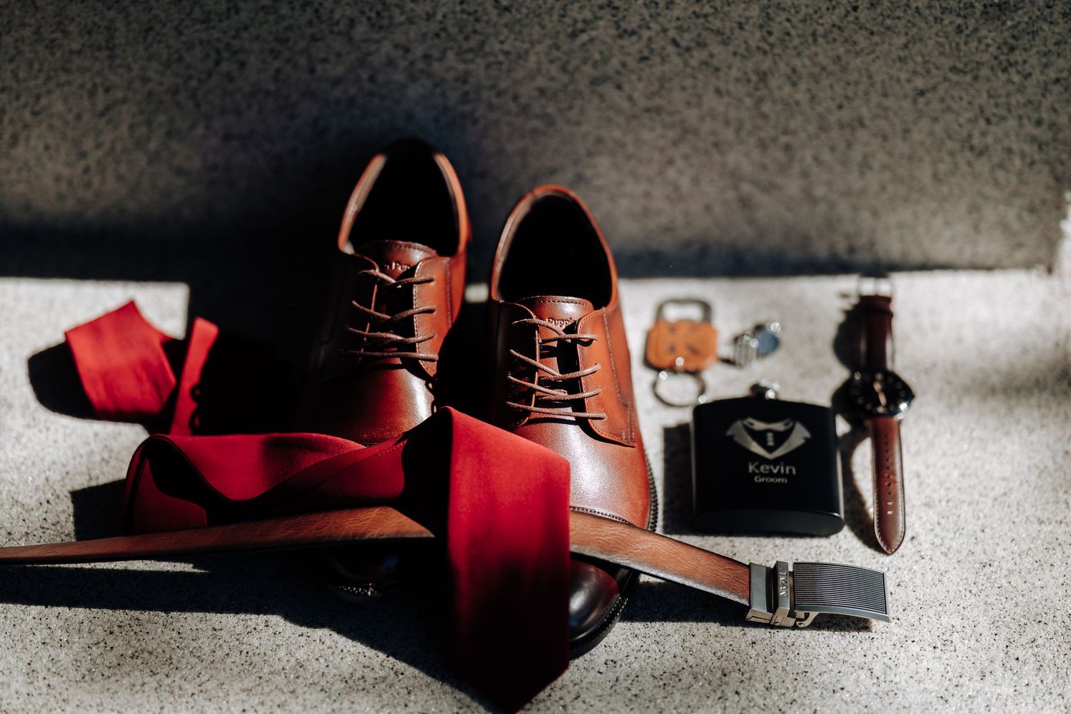 Brown leather Oxford shoes with red tie, belt, cologne, and accessories arranged on a gray surface.