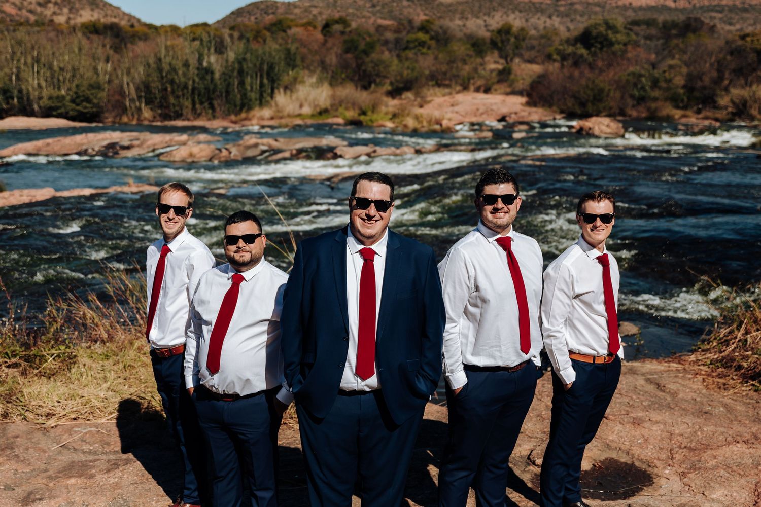 Five groomsmen in navy suits, white shirts, and red ties pose outdoors by a rocky river on a sunny wedding day.