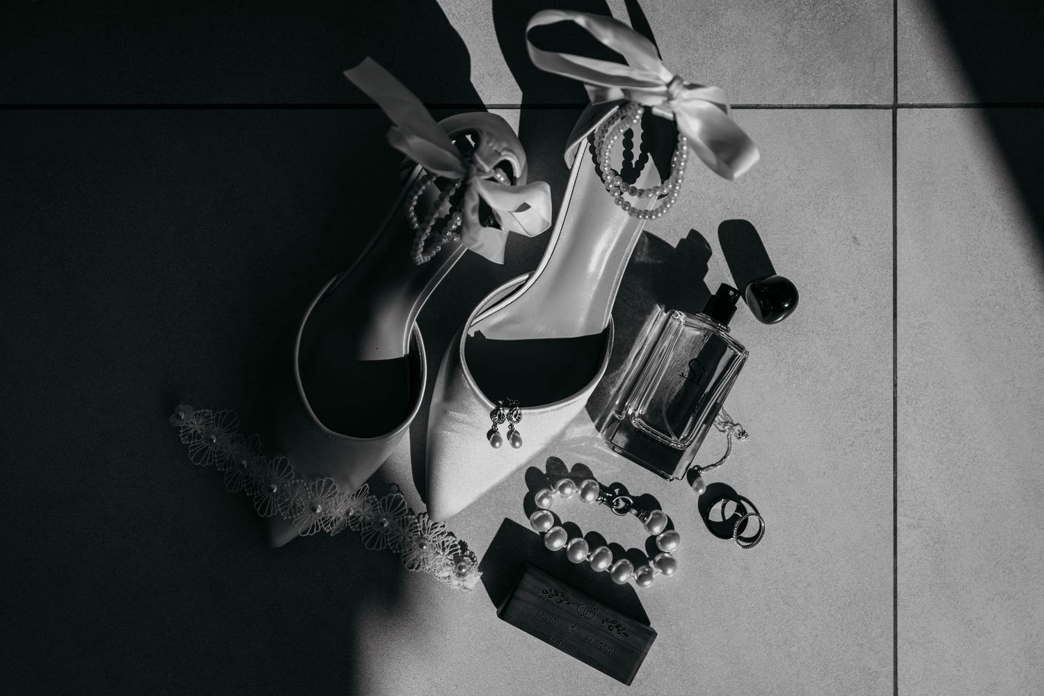 Black and white flat lay of bridal accessories including heels, perfume, jewelry, and a garter on a tiled floor.
