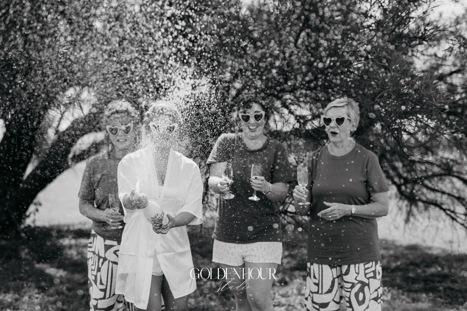 Four women celebrate outdoors with champagne in a joyful black and white photo.
