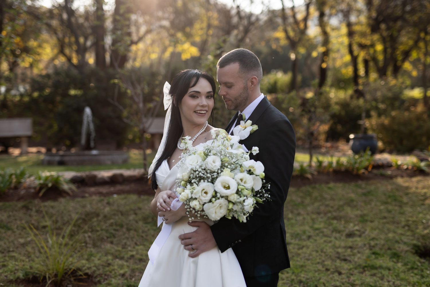 A couple in wedding attire stands close together in a garden setting while holding a white floral bouquet.