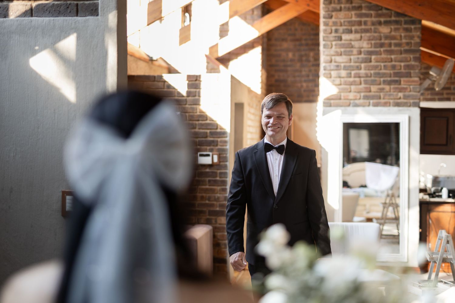A formal wedding scene at a rustic indoor venue with exposed brick walls and wooden beams, illuminated by natural light.