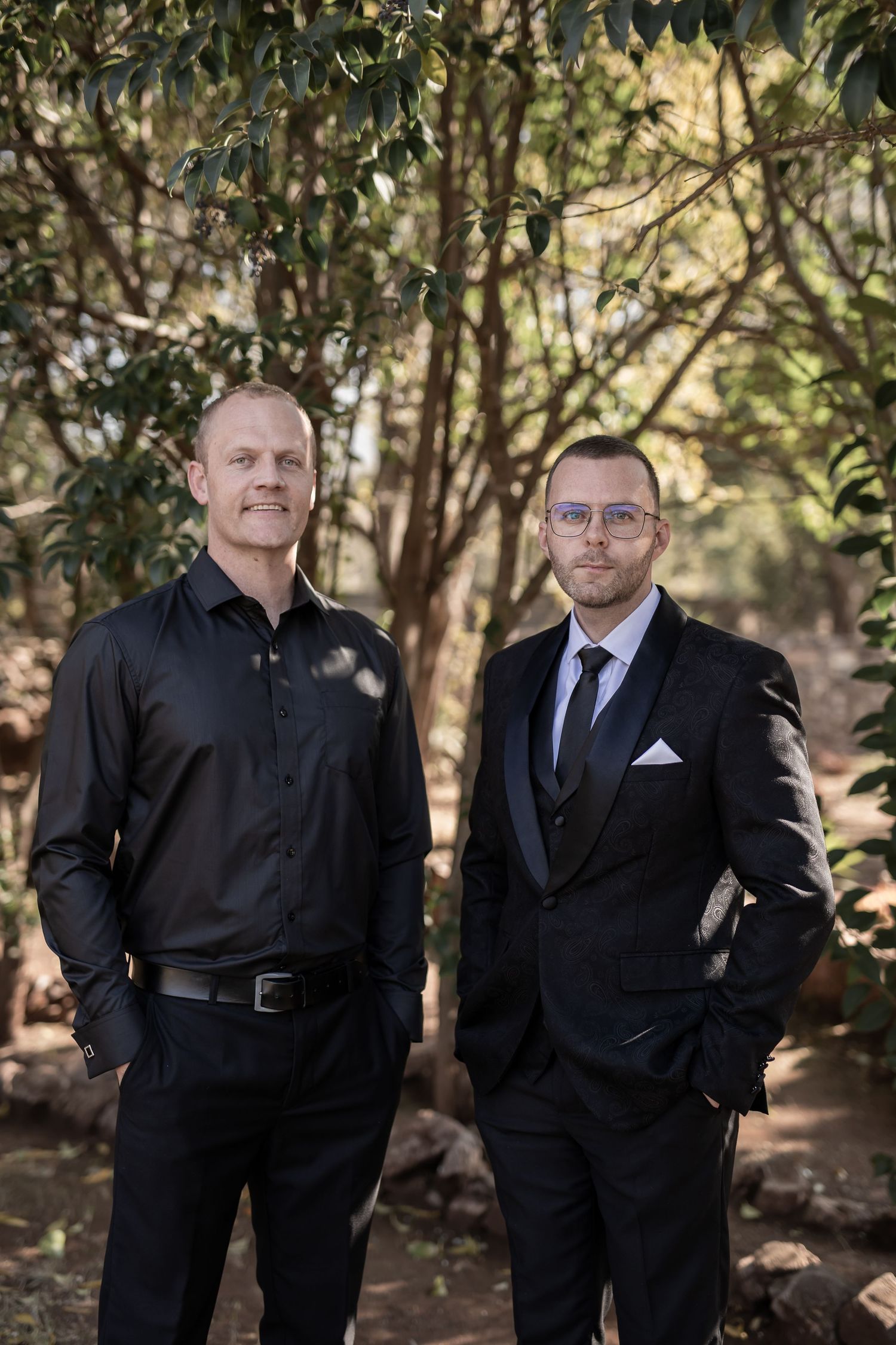 Two professionals in business attire standing together outdoors with trees and foliage in the background.