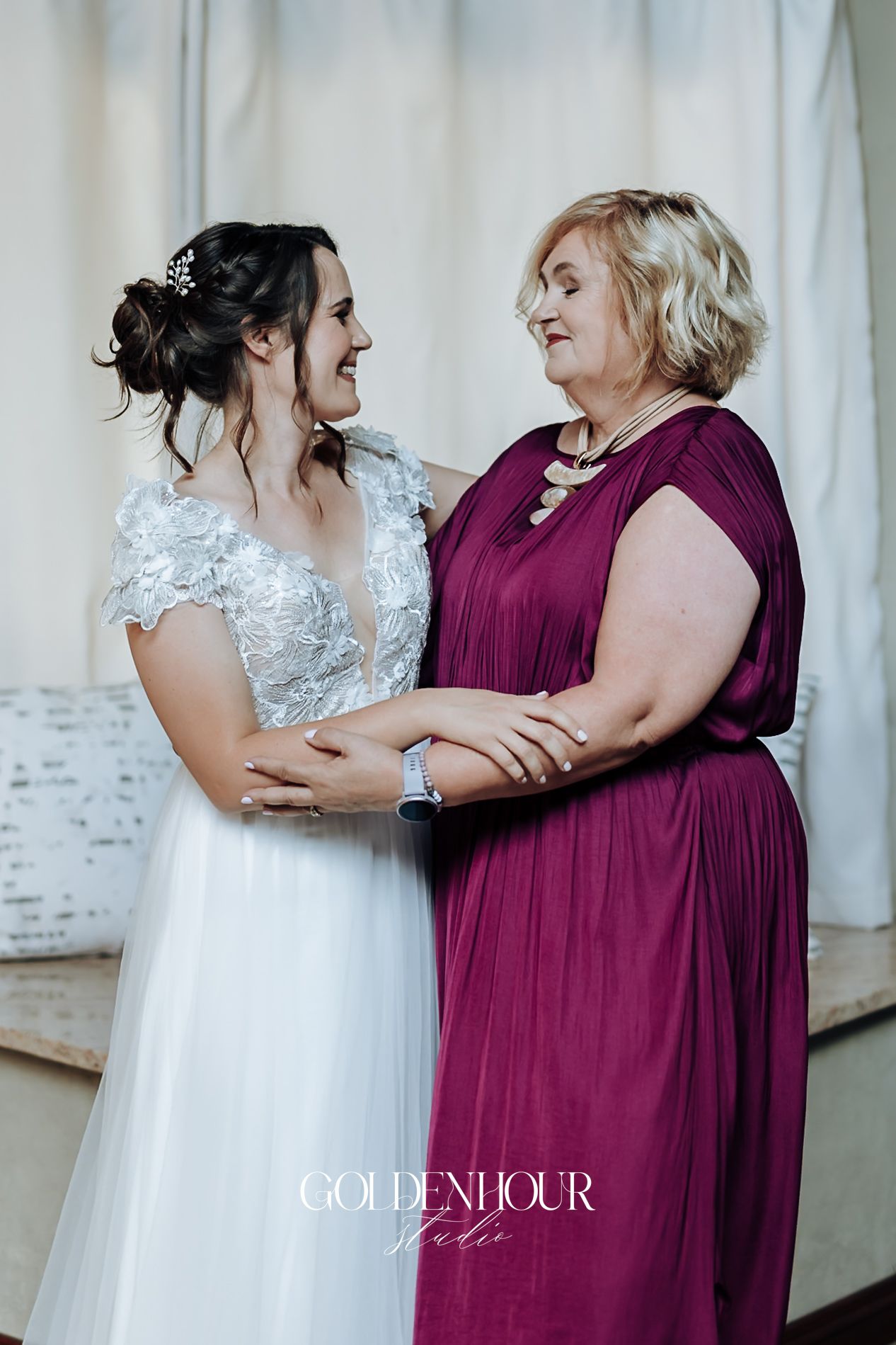 Bride in lace wedding gown smiling with mother in burgundy dress at wedding preparation.