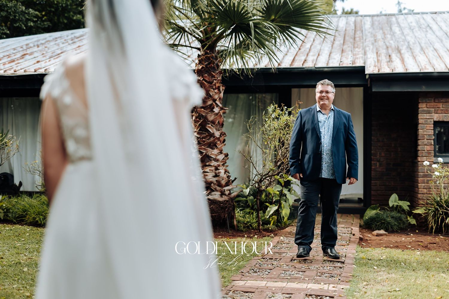 Father smiling as he approaches bride in white wedding gown outside a rustic building with palm tree.