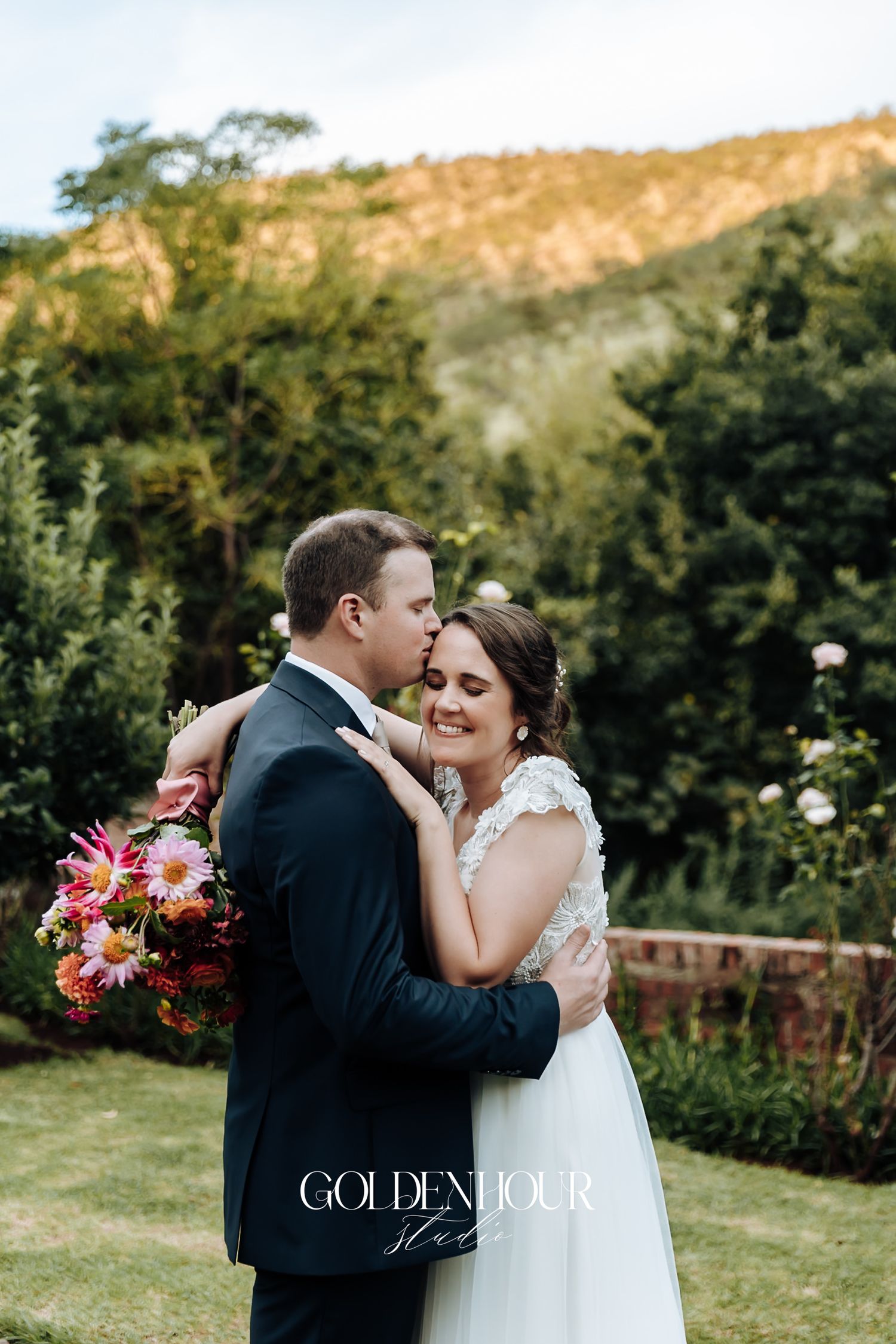 Groom tenderly kisses bride on forehead while embracing outdoors with mountain backdrop.