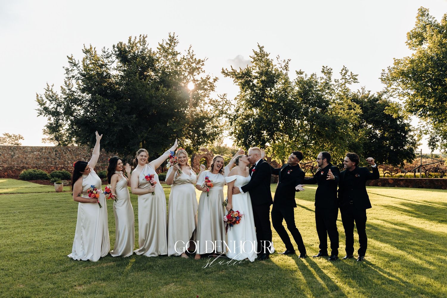 Wedding party stands together on a grassy hill during golden hour sunset creating a joyful outdoor celebration scene.