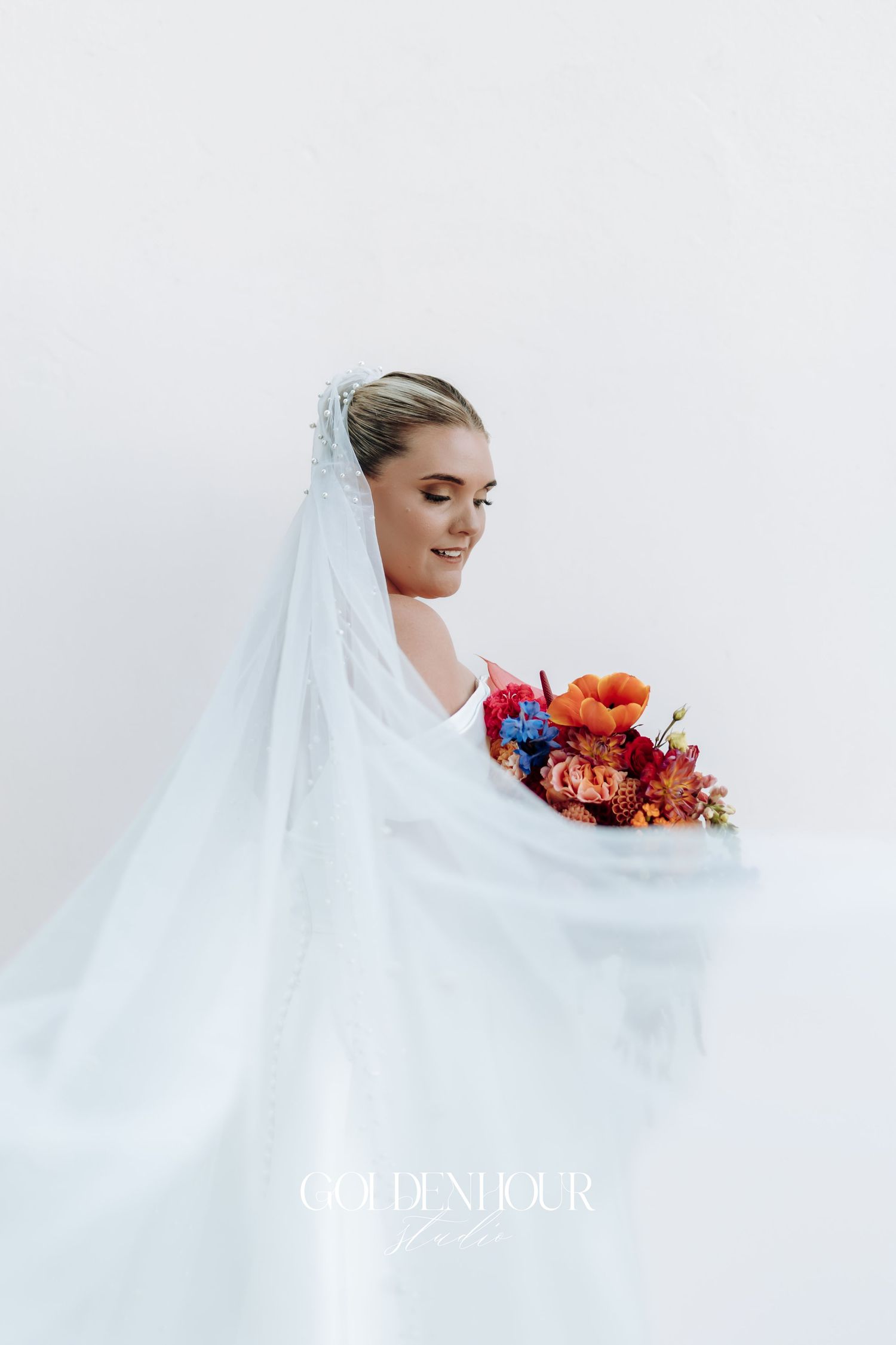 A bride in a flowing white veil poses with vibrant coral and pink flowers against a light background.