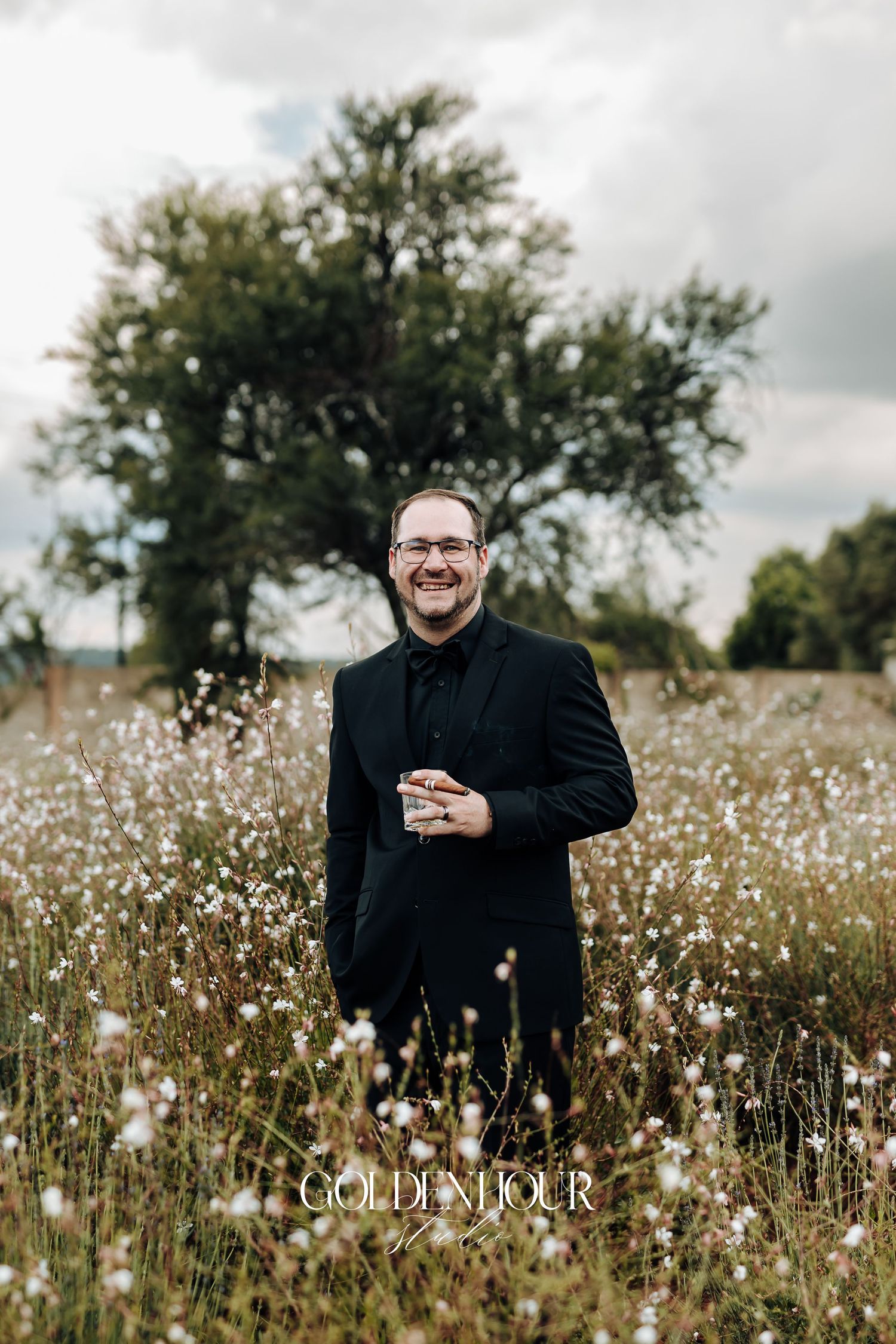 Person wearing black suit stands in a wildflower meadow with tall grass and white flowers under cloudy skies.