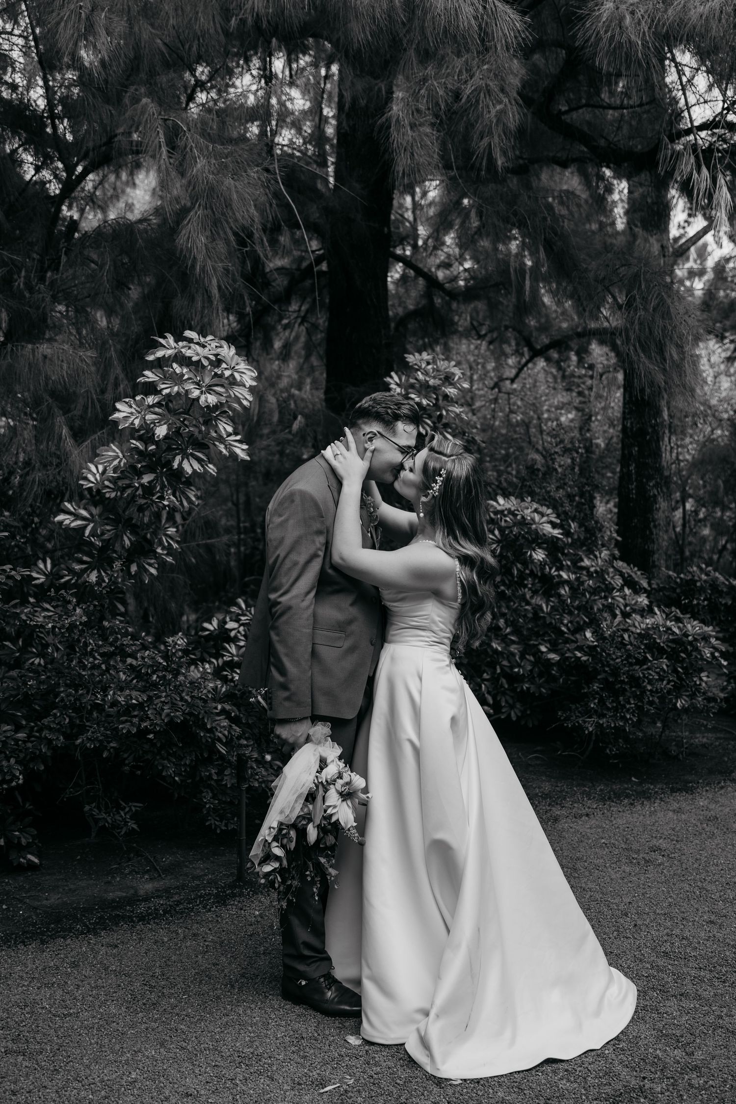 A romantic black and white wedding photo of a couple embracing outdoors in a garden setting with tall trees.