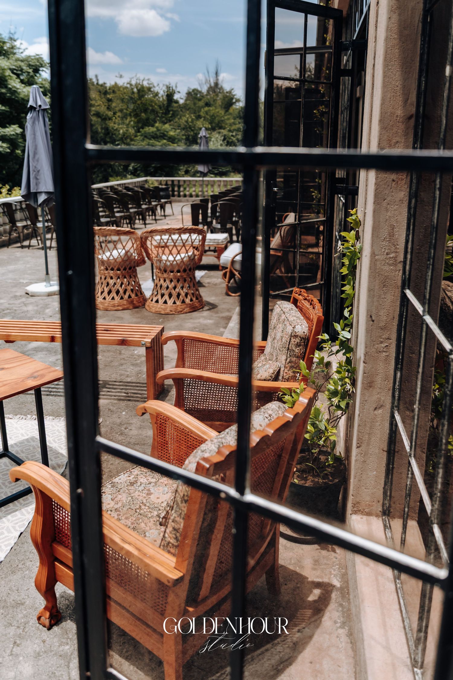 Vintage wicker chairs and wooden tables on an outdoor patio viewed through black metal window bars.