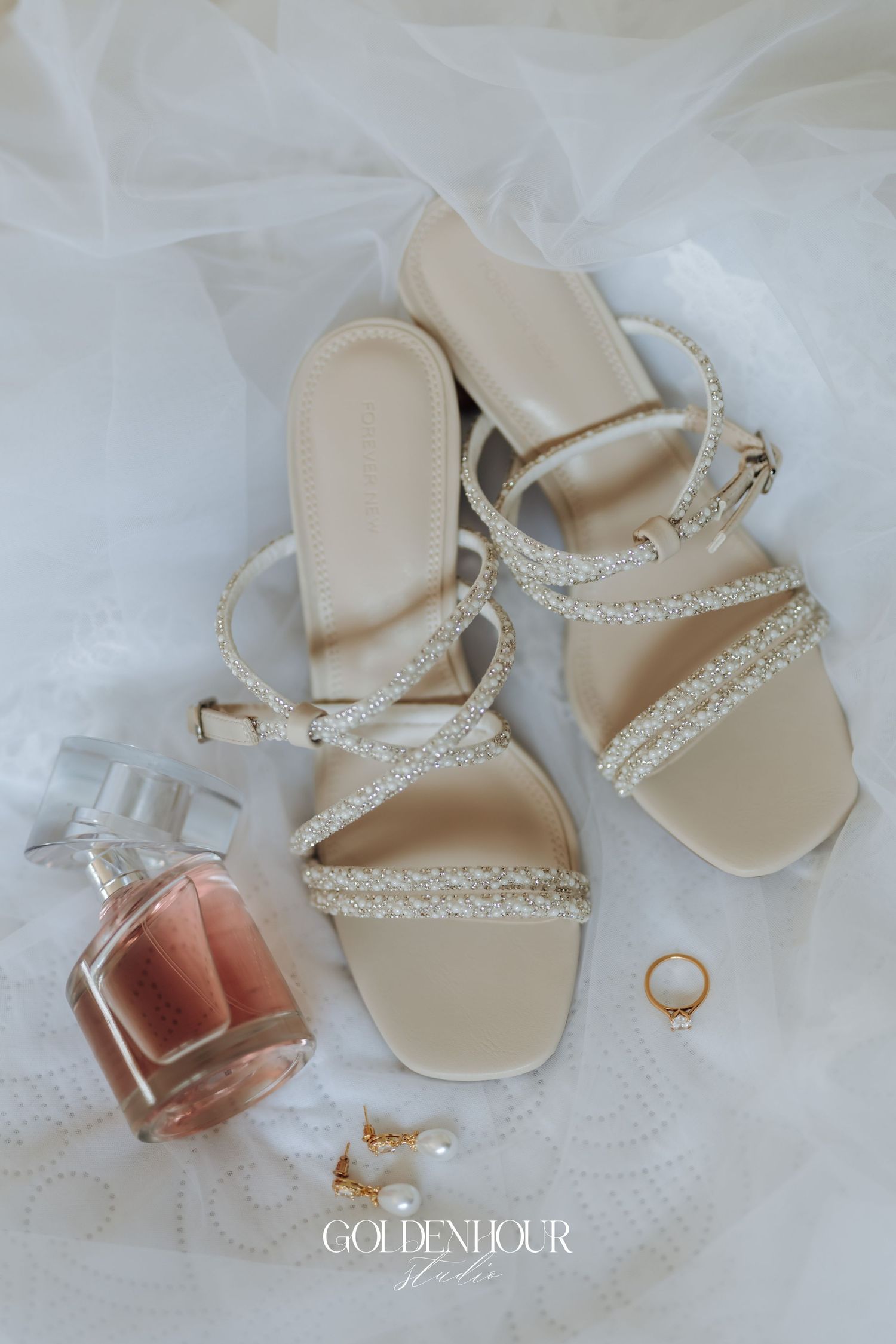 Wedding accessories including elegant beige sandals, perfume bottle, and rings arranged on white fabric.