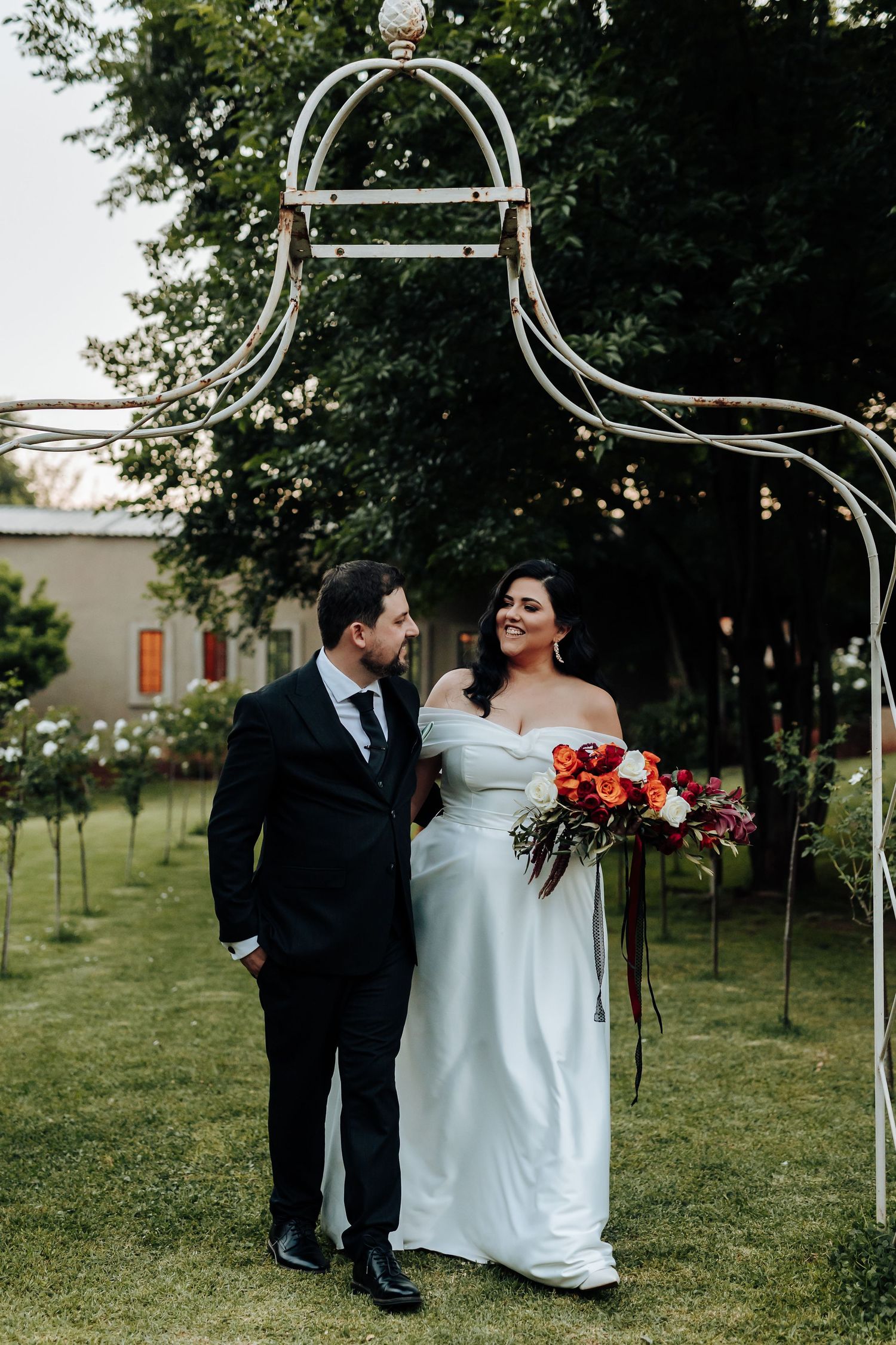 A wedding couple shares a tender moment under a decorative arch in a garden setting.