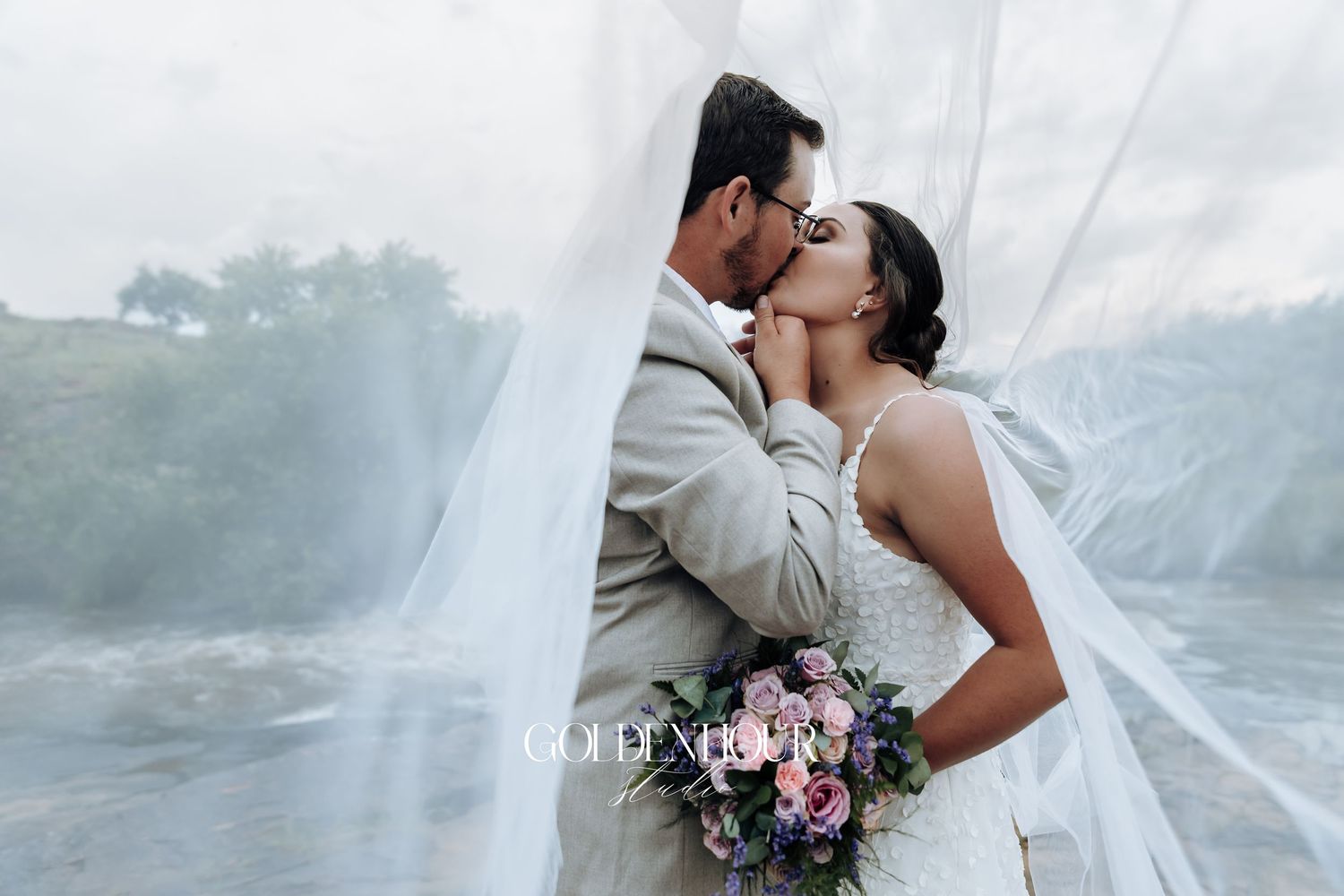 Wedding couple embraces as veil flows in the wind against cloudy sky.