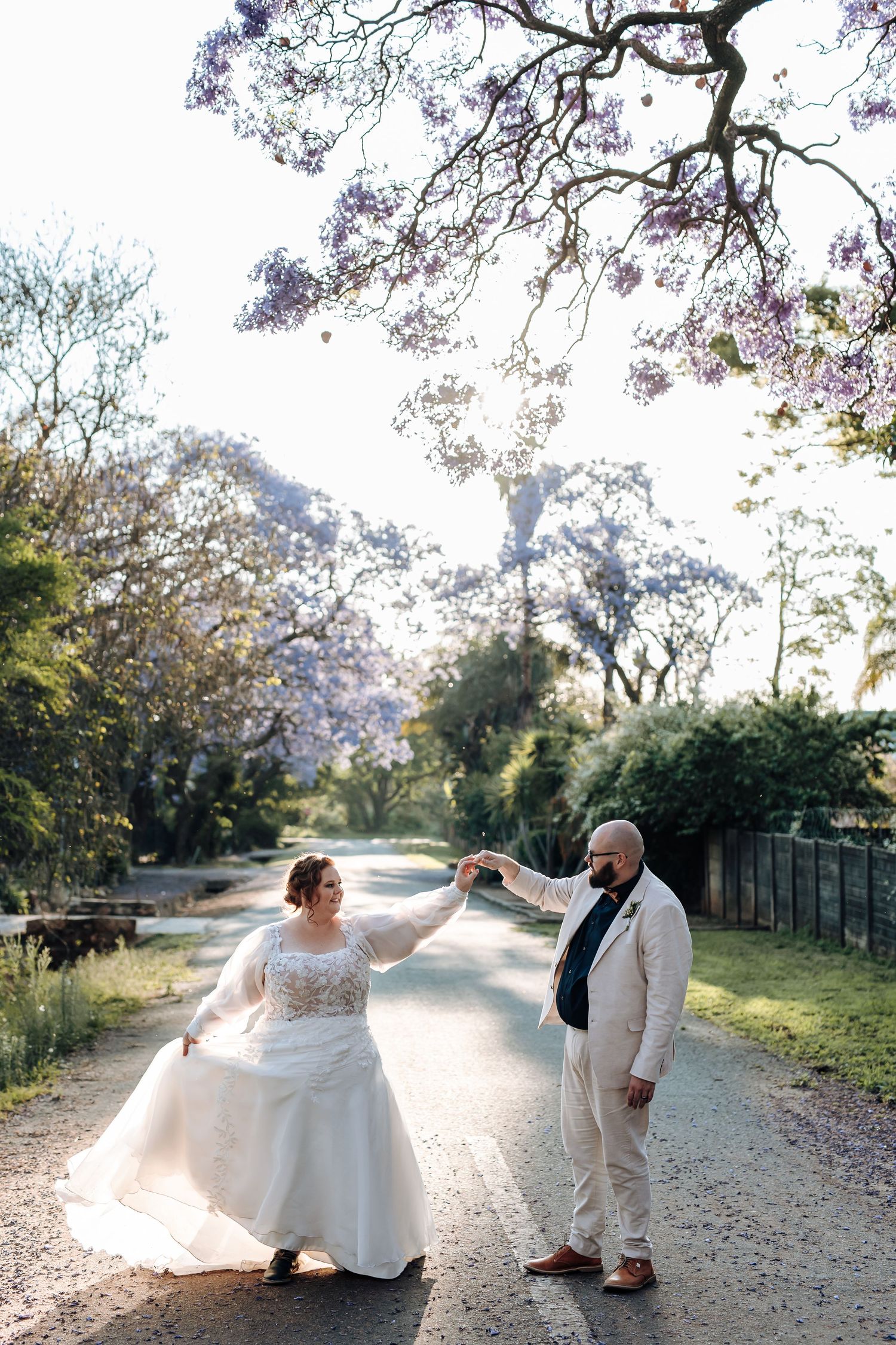 A couple in wedding attire dance together on a tree-lined path during a romantic outdoor photo session.