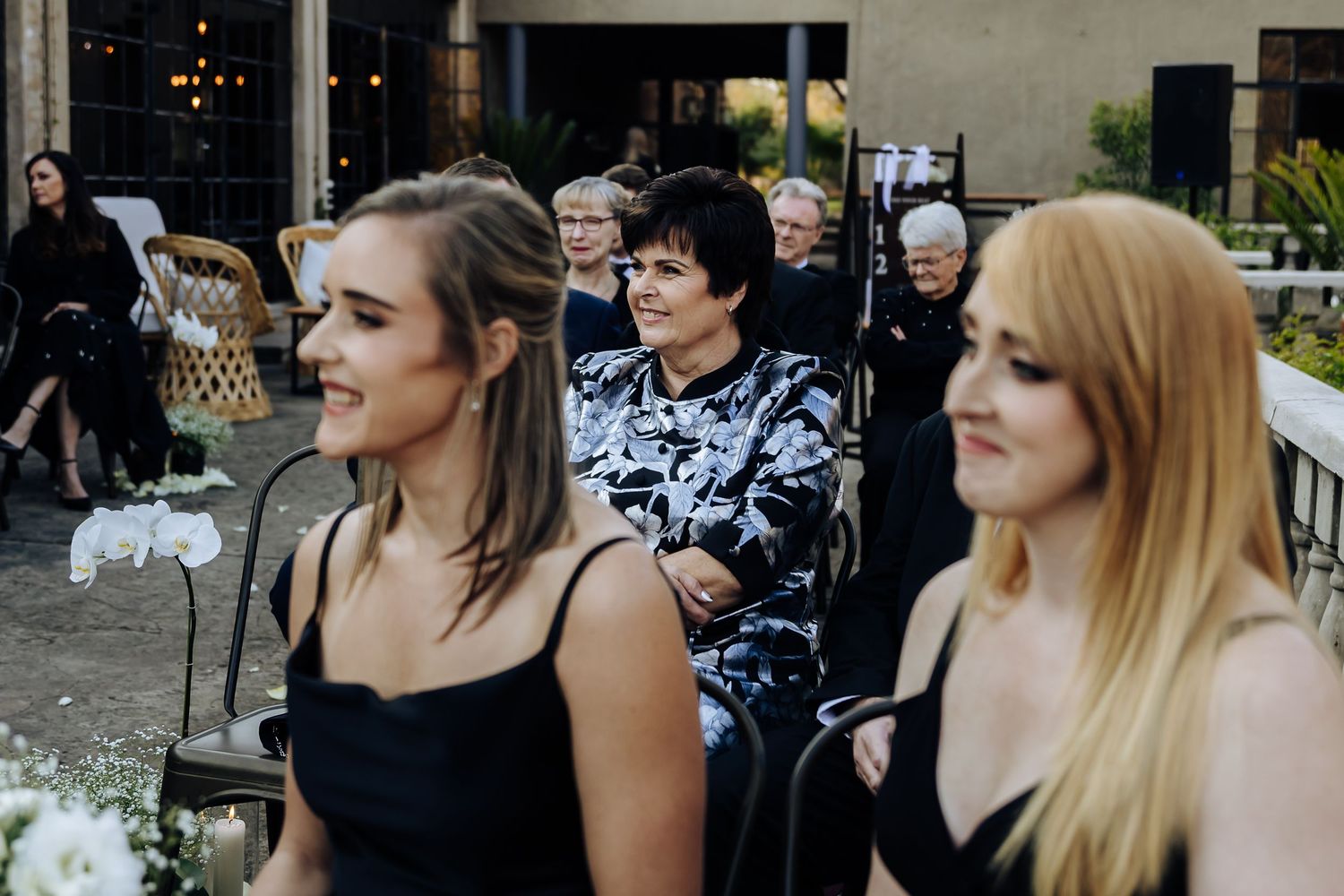 Party guests in dark formal attire sitting at an outdoor evening reception with decorative lighting.