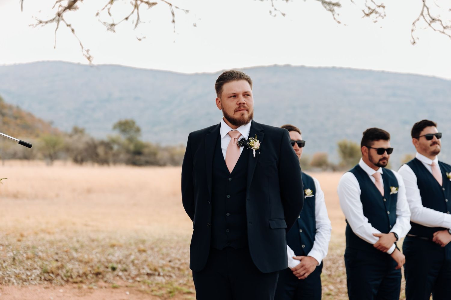 A formal wedding party of groomsmen stands together in dark suits against a scenic mountain backdrop in desert terrain.