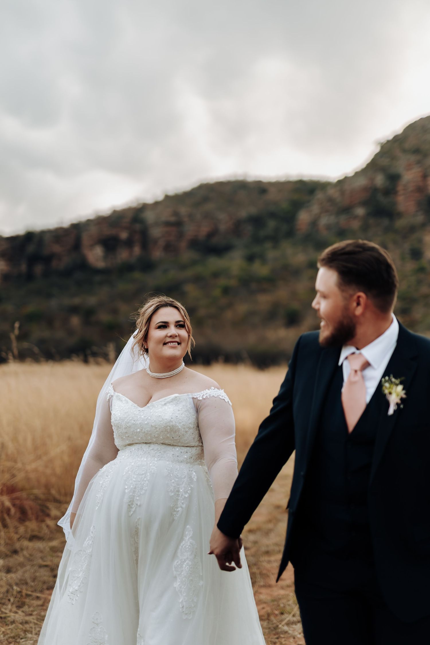 A couple in wedding attire walks through a golden field with mountains in the background as the sun sets.