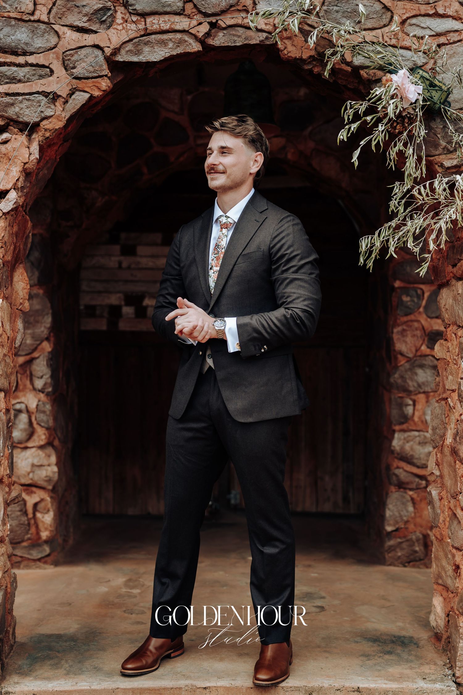 Person in a dark suit and brown shoes poses in a rustic stone archway setting.
