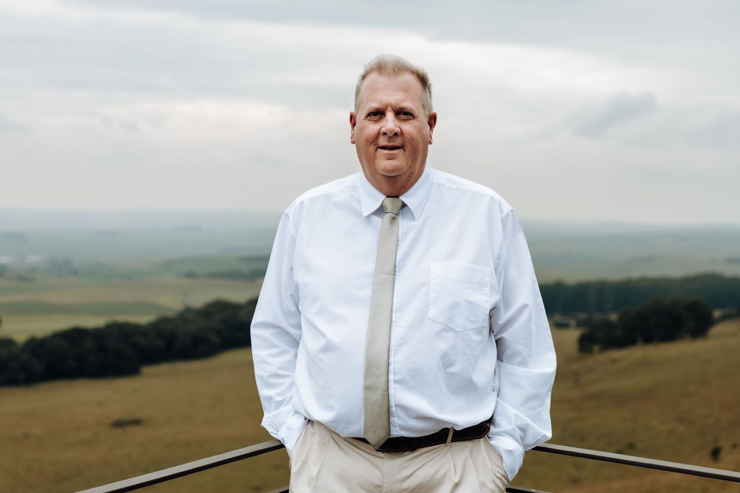 A person in a white dress shirt and tie stands on a hilltop overlooking rolling countryside under cloudy skies.