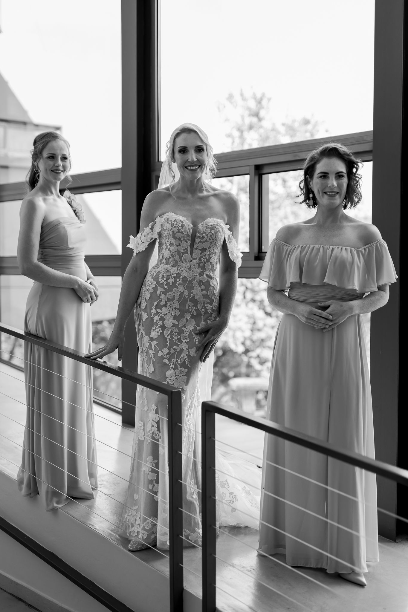 Three women in formal dresses pose on indoor balcony with glass railing.