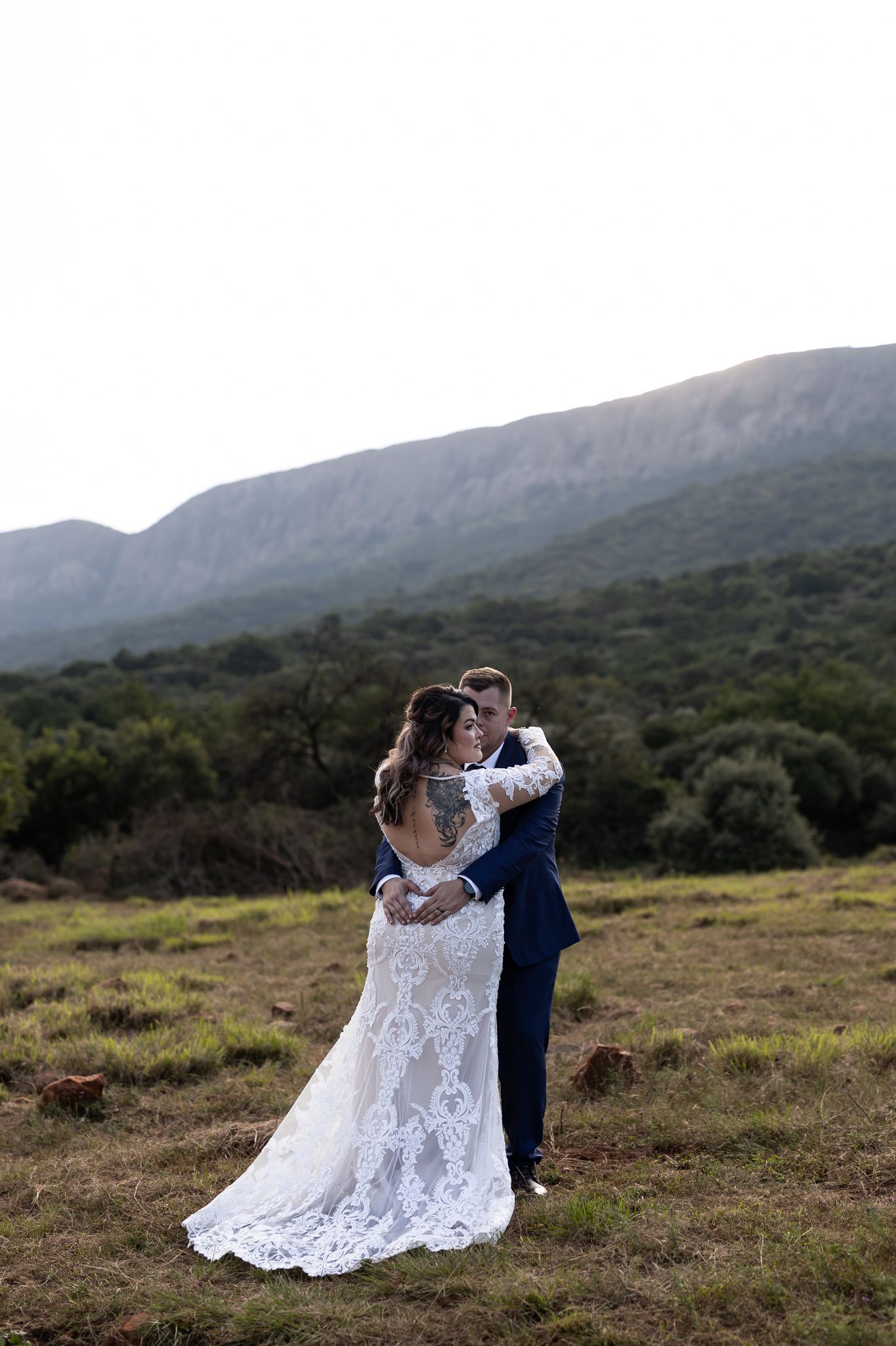 A couple in wedding attire embraces against a mountain backdrop in a rustic outdoor setting.