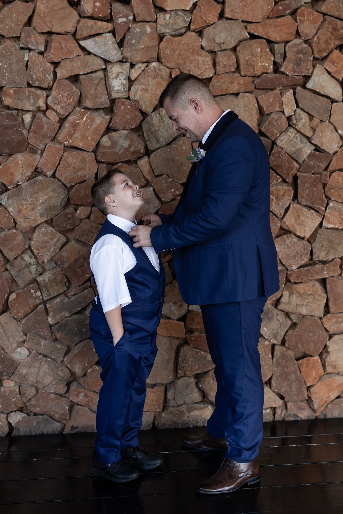 Two people in navy and white formalwear share a moment against a stone wall backdrop.