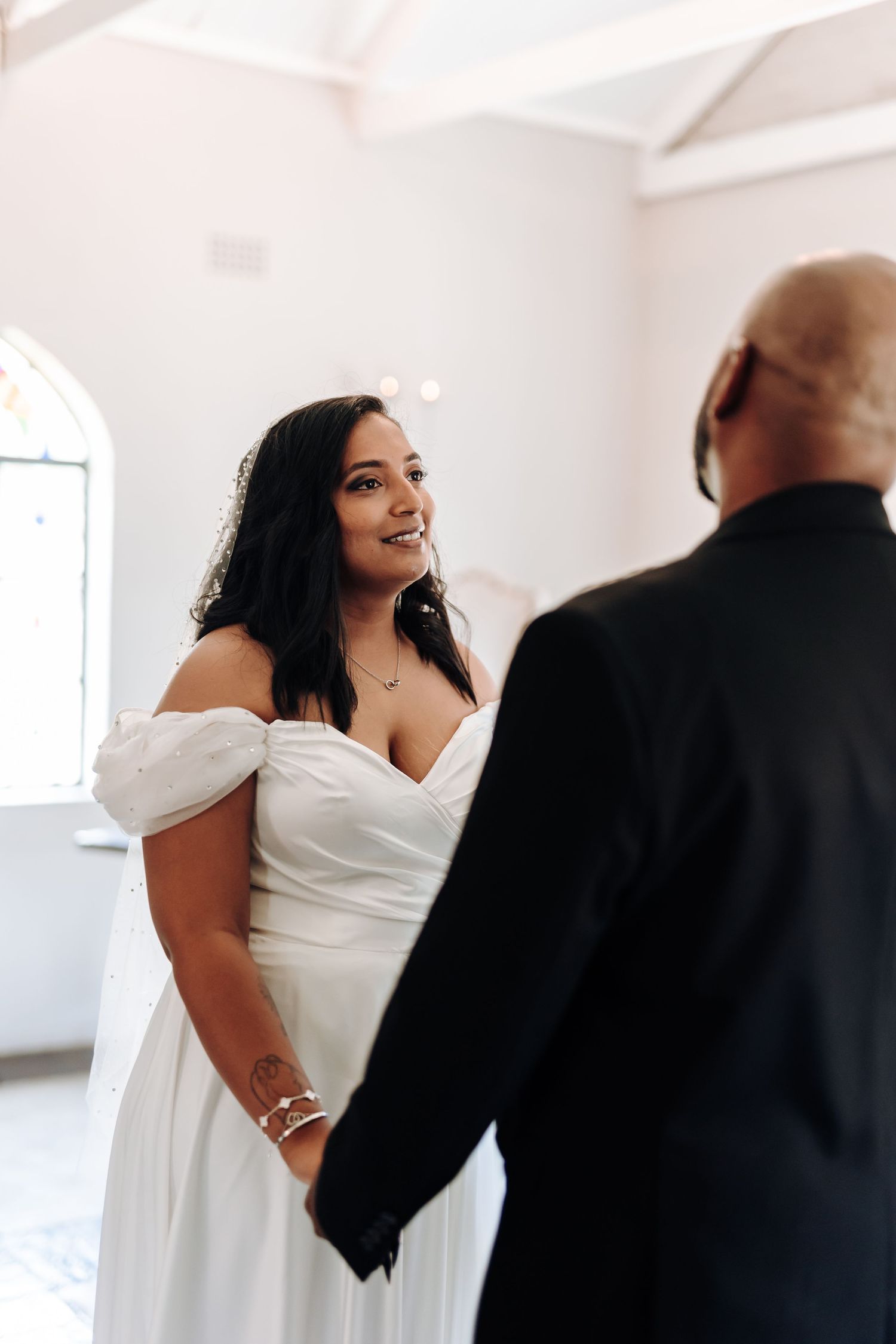 A bride in an off-shoulder white gown smiles during her wedding ceremony.