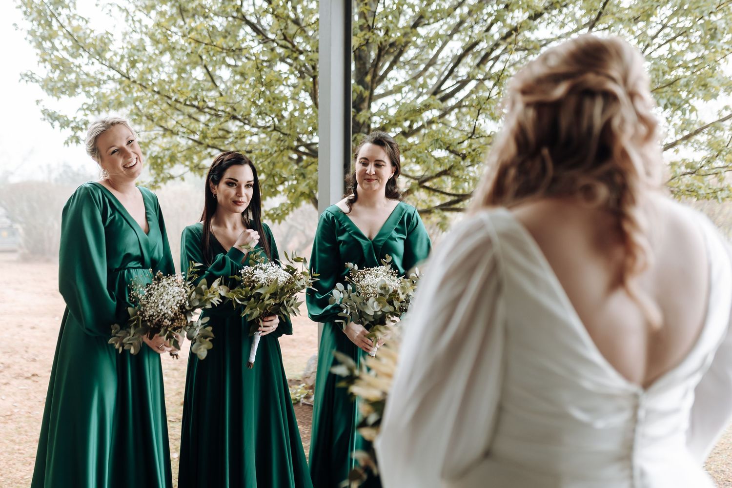 Wedding party in emerald green dresses with dried flower bouquets gathering outside.