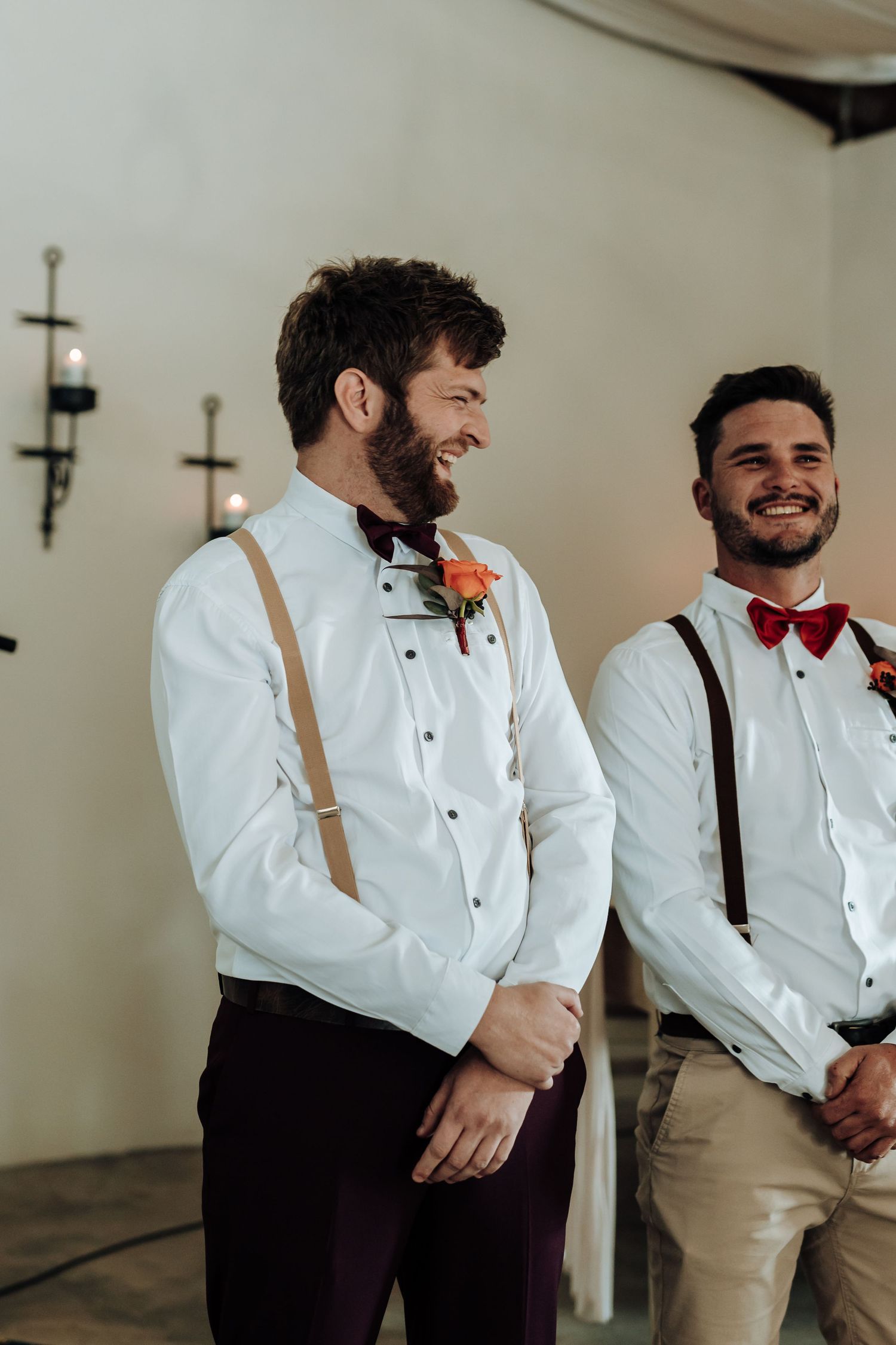 Two groomsmen in white shirts with suspenders and red bowties share a candid moment.