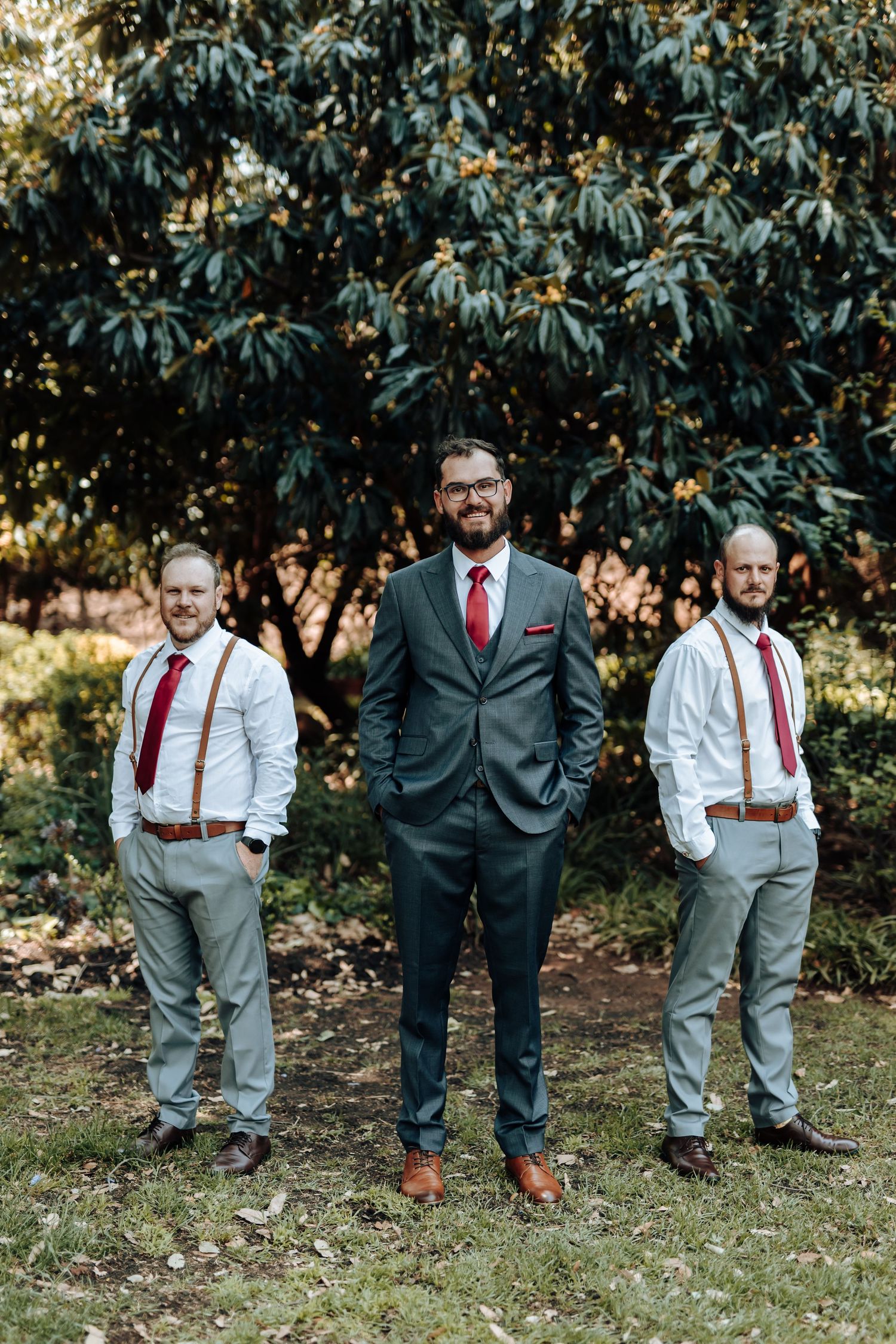 Three groomsmen pose in coordinating formal attire with red ties against a lush garden background.