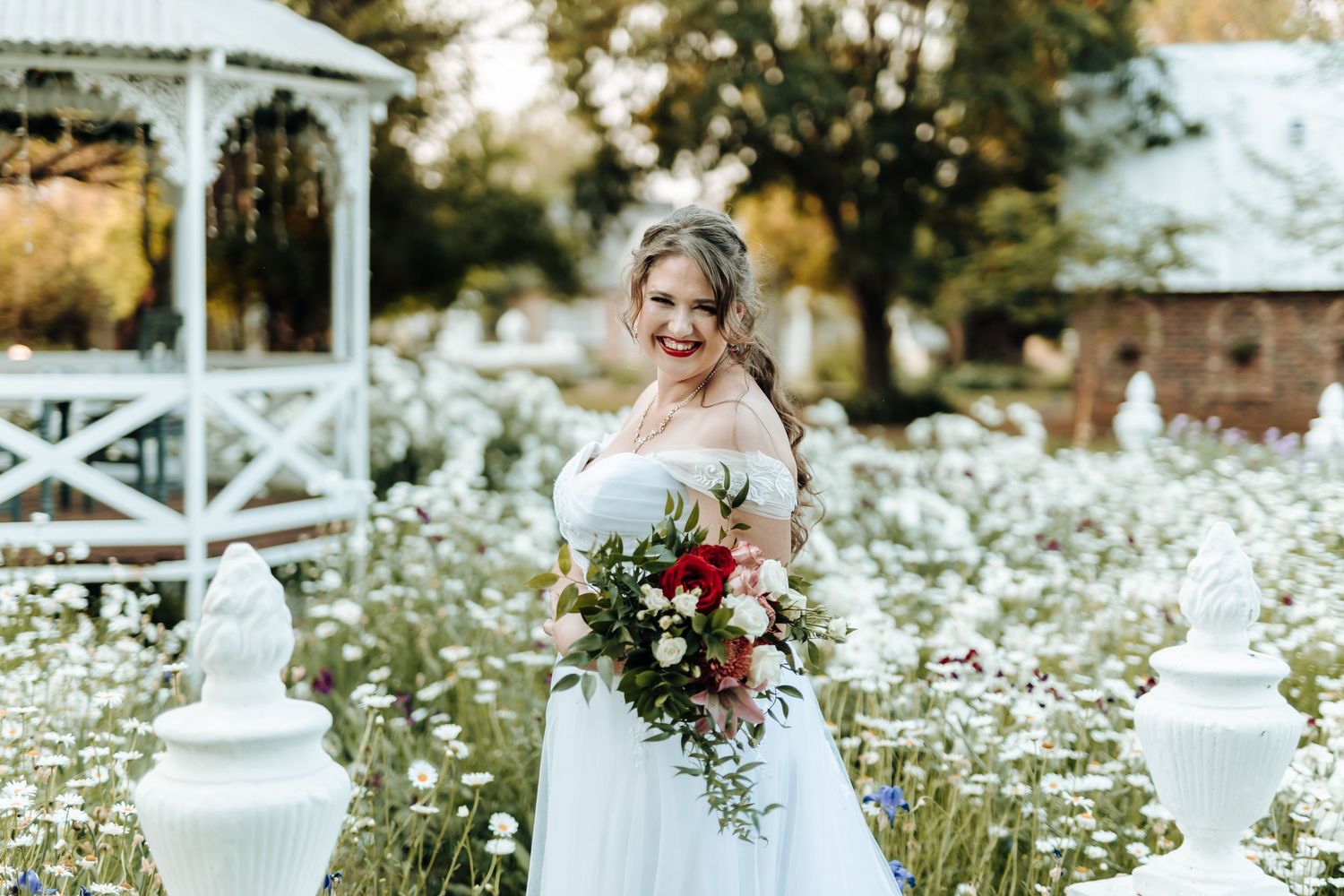 Wedding portrait in a garden with white gazebo and wildflowers featuring elegant white gown and red bouquet.