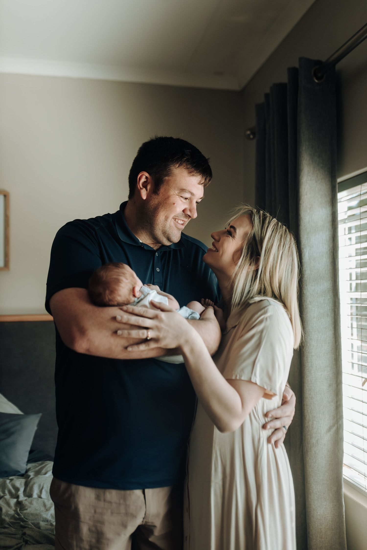 A couple shares a tender embrace while standing near a sunlit window in a cozy room.