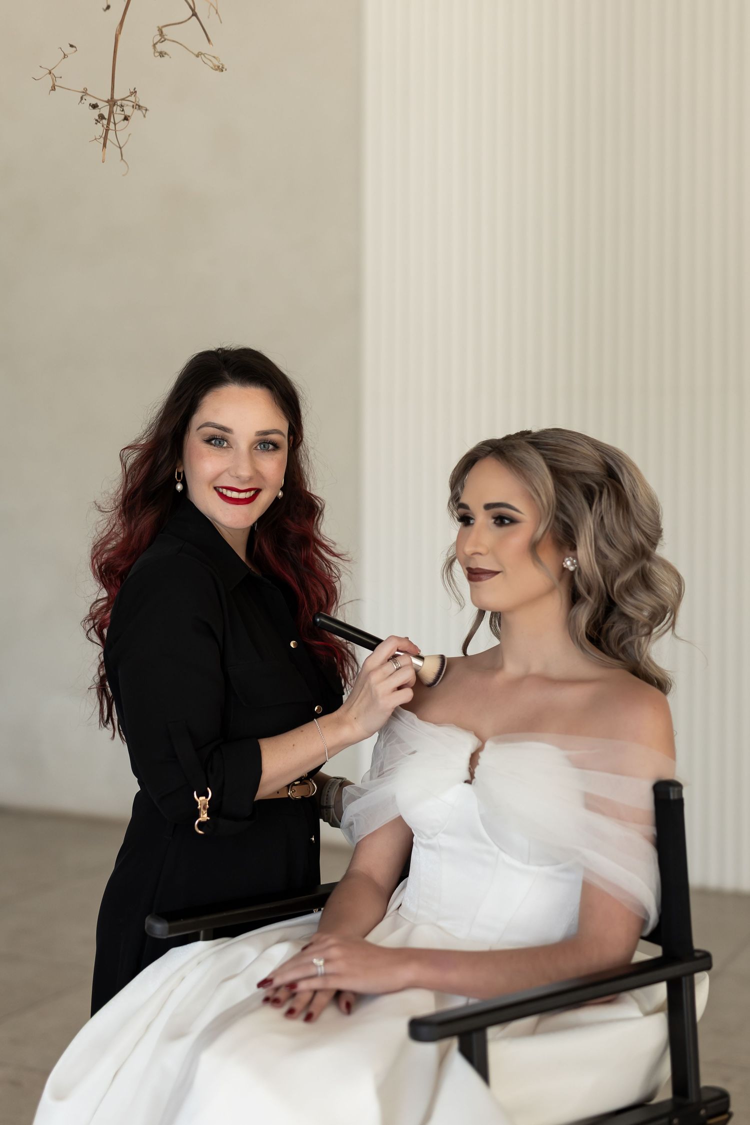 A makeup artist in black applies cosmetics to a bride wearing a white strapless gown.