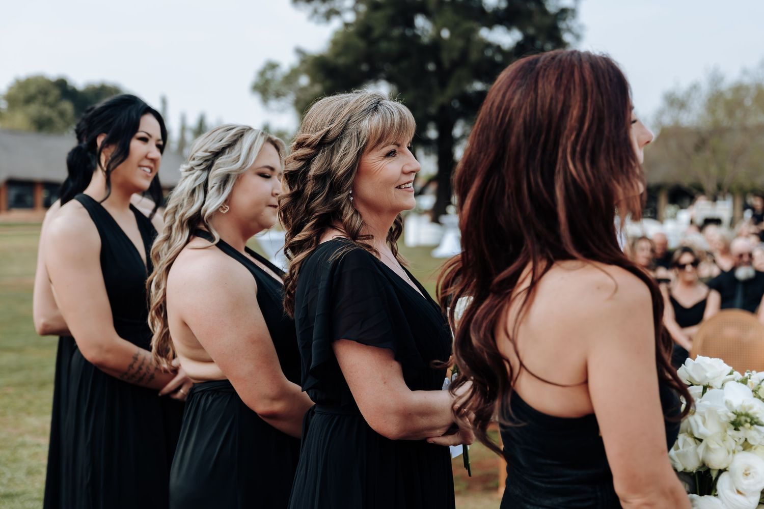 A group of bridesmaids in black dresses share laughter and hugs during an outdoor wedding celebration.